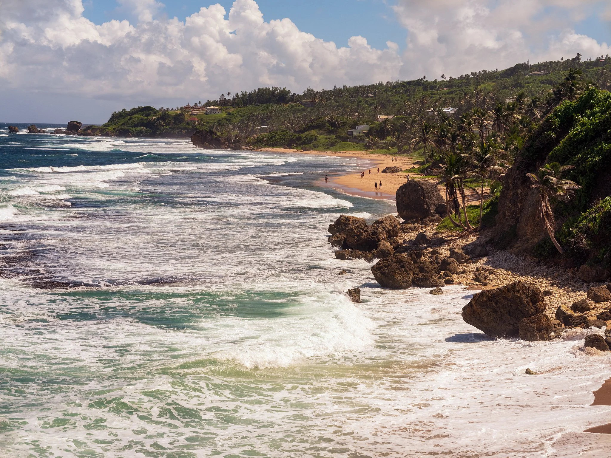 A scenic view of a tropical beach with ocean waves crashing onto the sandy shore, rocky outcroppings, and lush greenery with palm trees, houses on hills, and a partly cloudy sky.