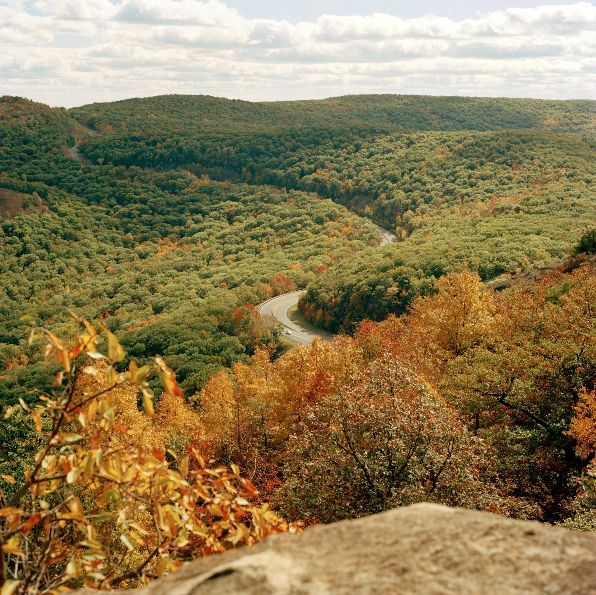 A scenic view of a winding road through a forest upstate New York with trees displaying fall colors, hills in the background, and a partly cloudy sky.