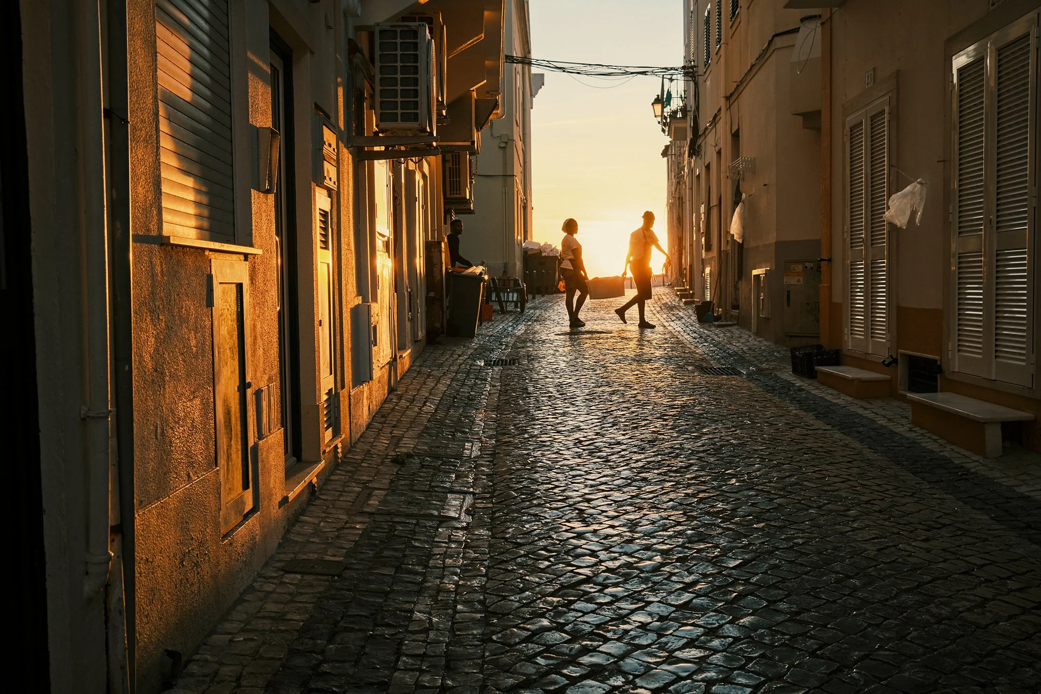 Silhouettes of two women carrying a crate on a cobblestone street in Nazare in Portugal during sunset, with buildings on either side.