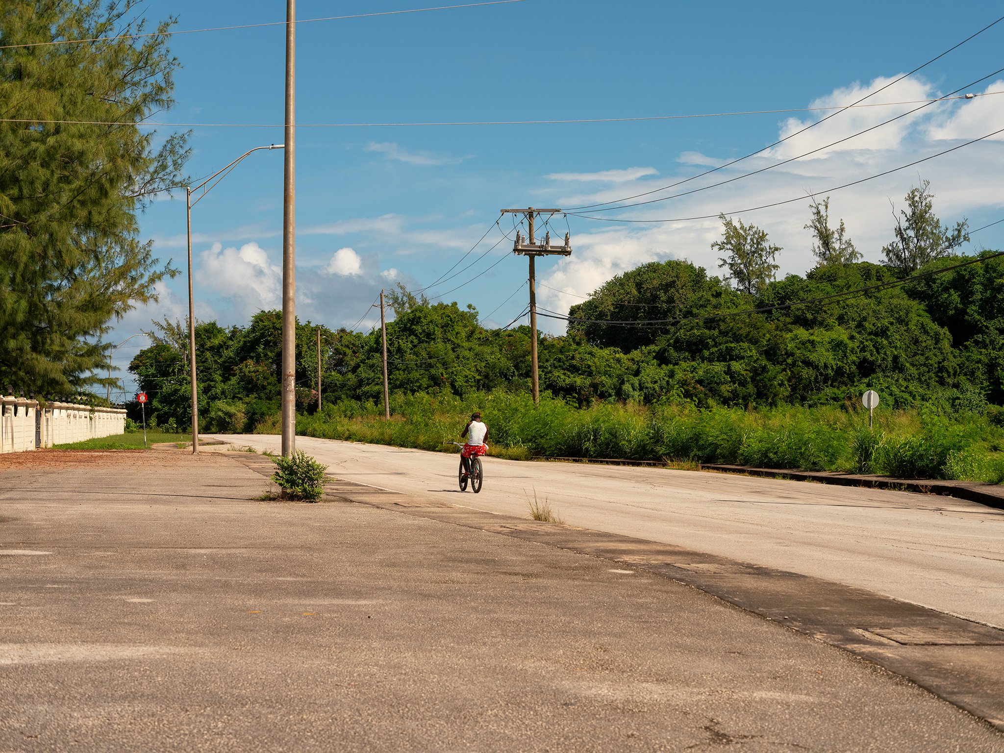 A teen riding a bicycle in Barbados on an empty street near green trees and utility poles under a partly cloudy sky.