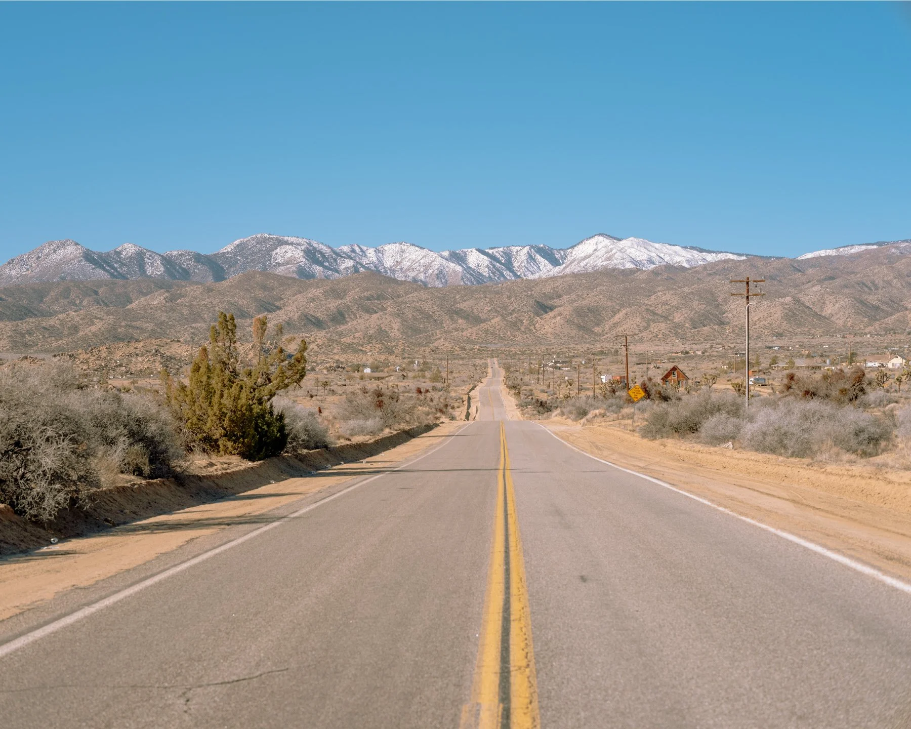 A straight empty road in a California desert landscape with mountains in the background and clear blue sky.