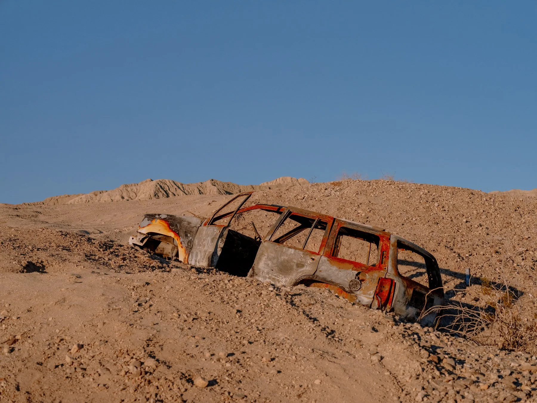 A burnt-out, abandoned vehicle in California partially buried in desert sand under a clear blue sky.
