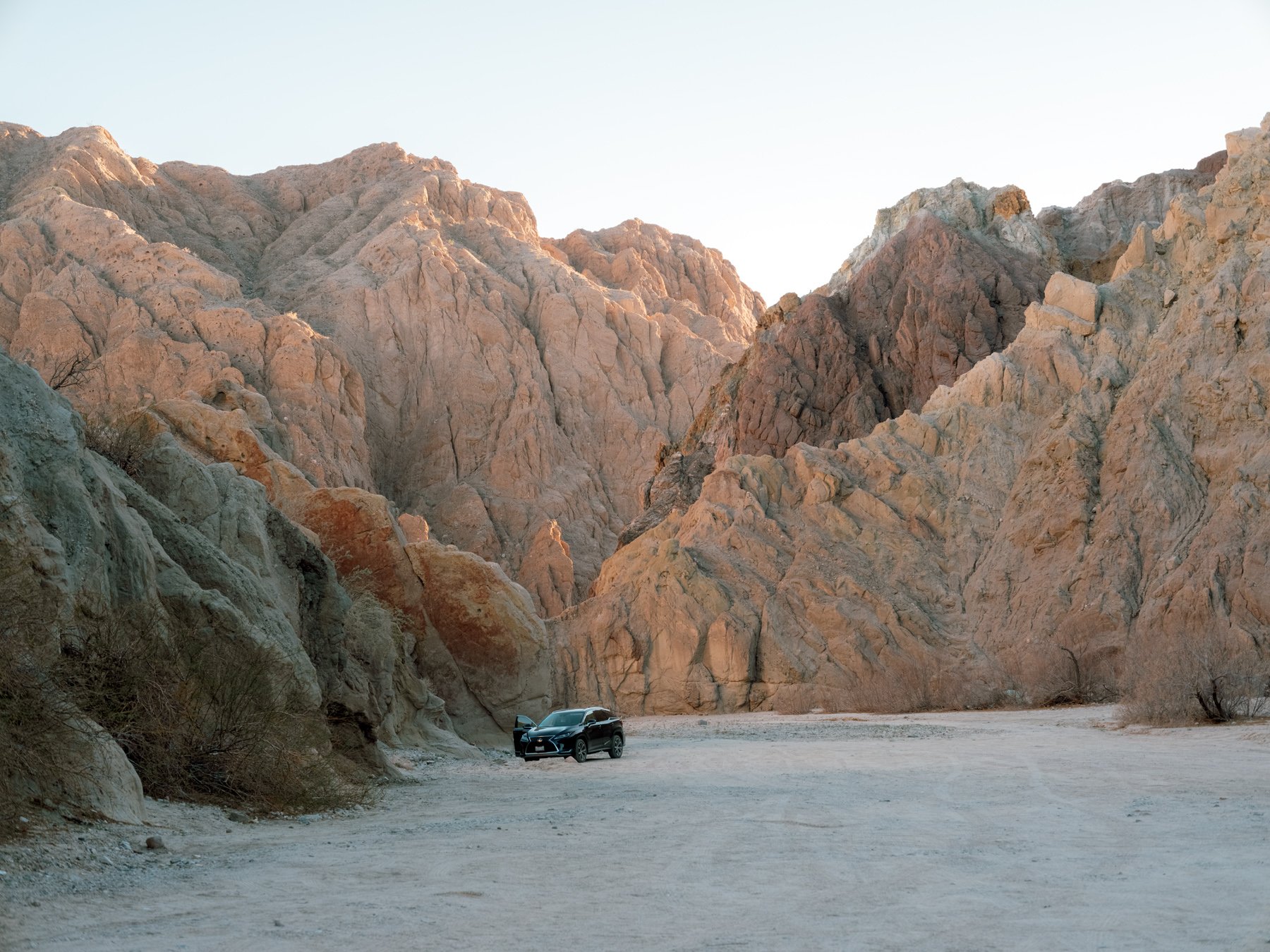 A dark-colored car parked on a flat, dry, arid landscape with large, rugged rocky mountains in the background, under a clear sky.