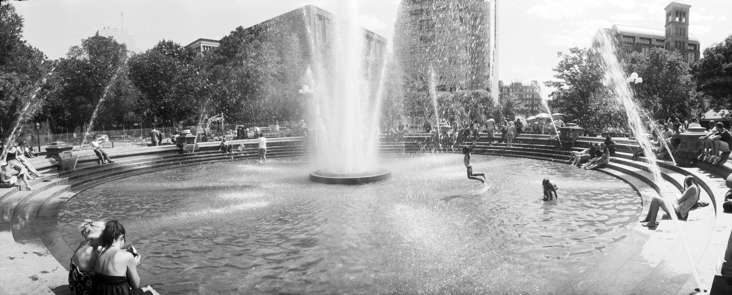 People enjoying a circular fountain with water jets in a city park, surrounded by trees and tall buildings in the background.