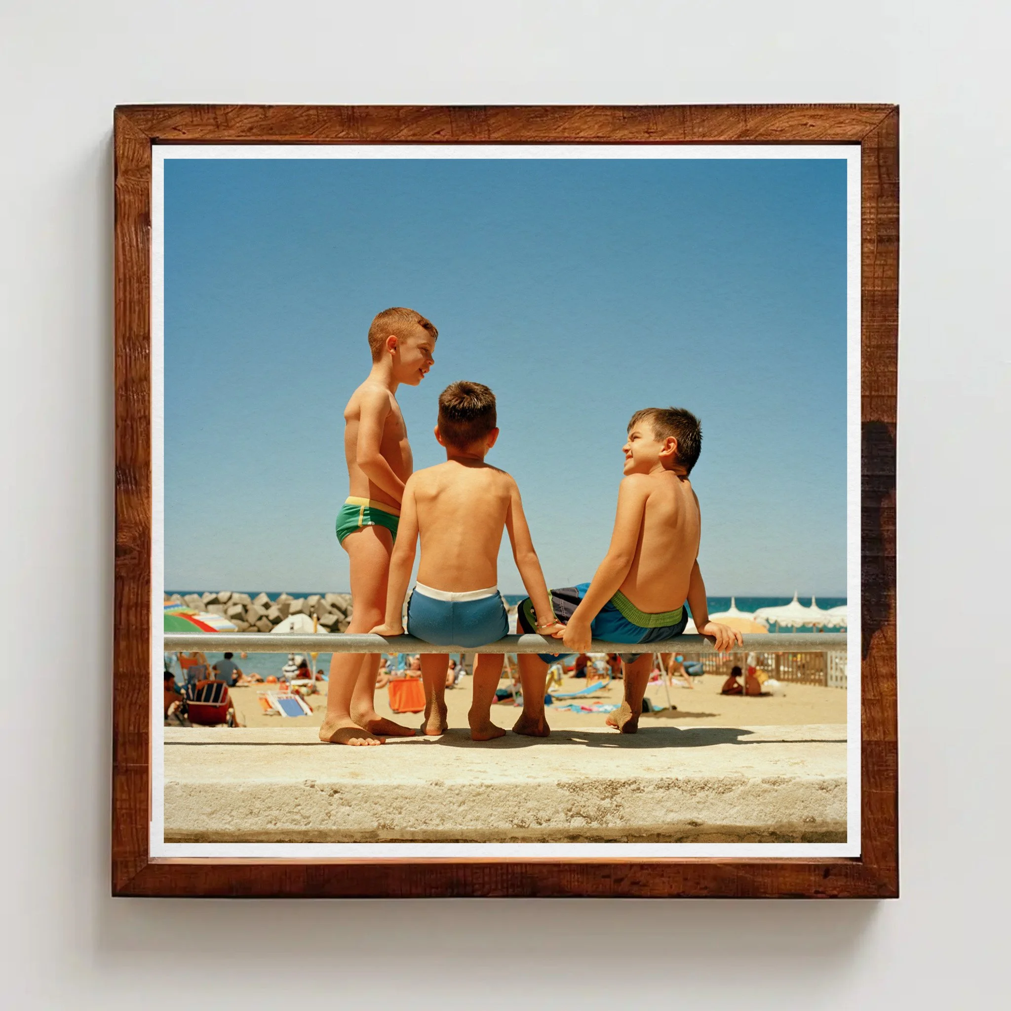 Three young boys in swimsuits sitting and talking on a rail at the beach with umbrellas and people in the background, under a clear blue sky.