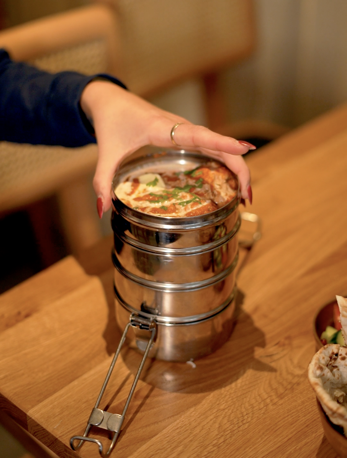 A hand holding an open metal Tiffin filled with freshly prepared curry at the restaurant