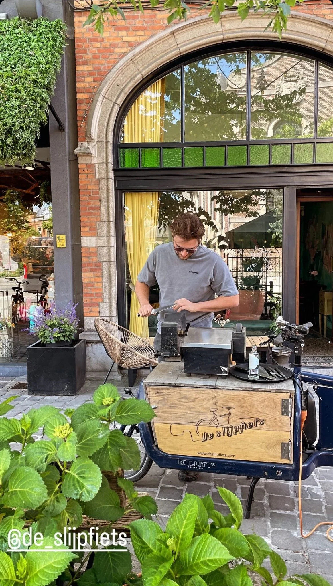 A man in sunglasses preparing food at a street food cart in front of a building with large windows and a brick facade, surrounded by greenery.