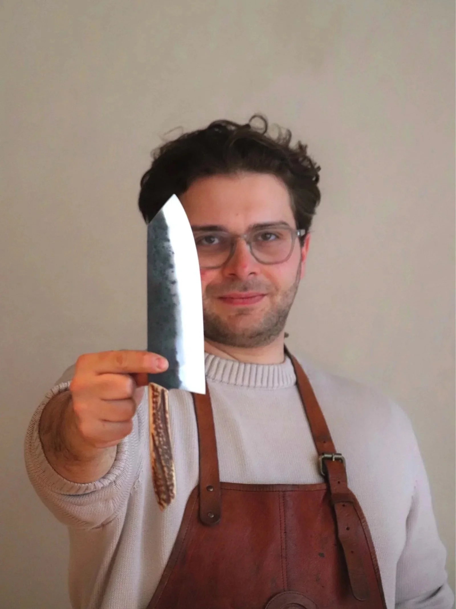 A man with dark hair, glasses, and a light-colored sweater wearing a brown apron, holding a large knife, standing against a plain background.