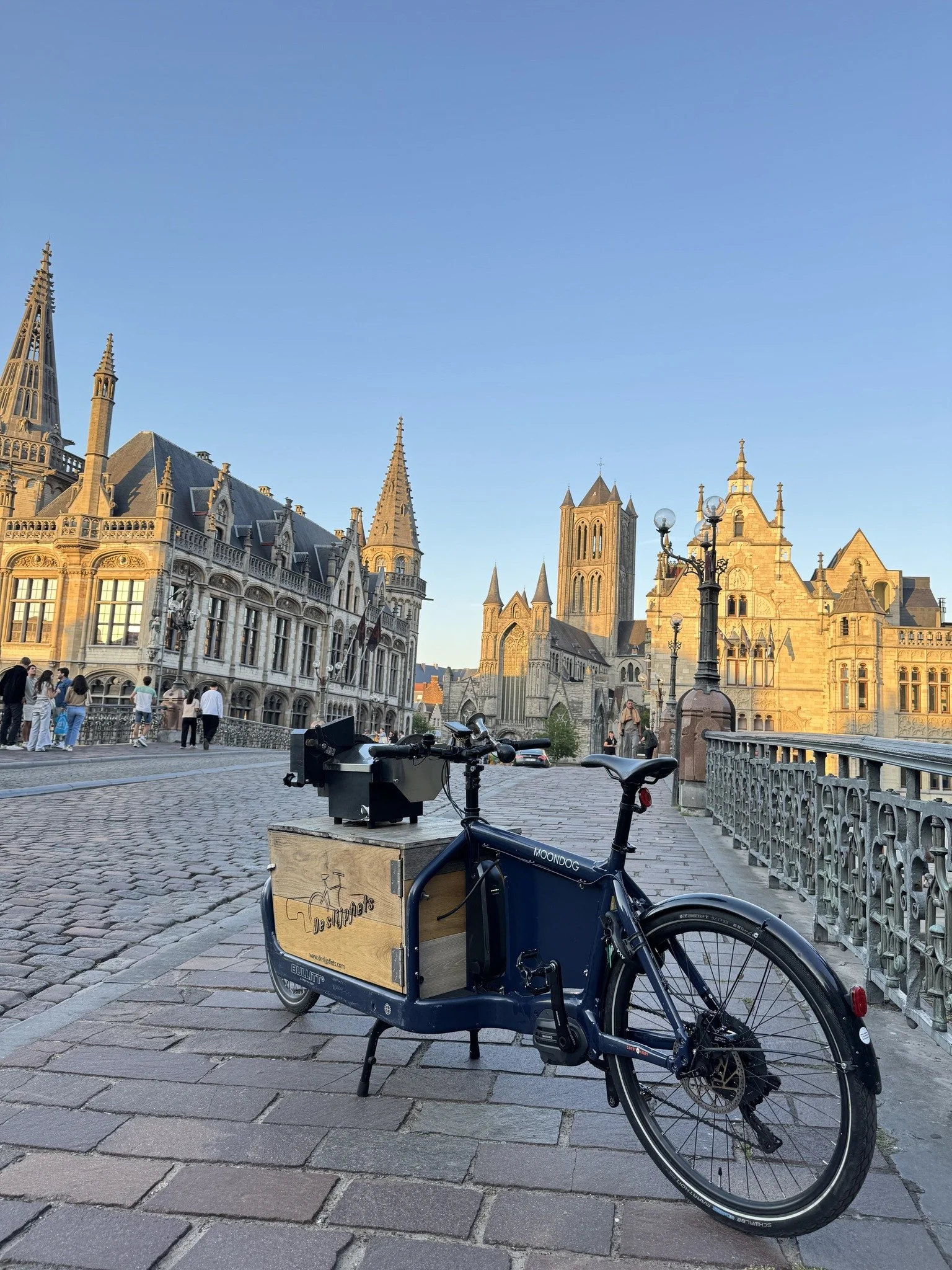 A blue cargo bike parked on a stone-paved street with historic buildings and a cathedral in the background, during the daytime.