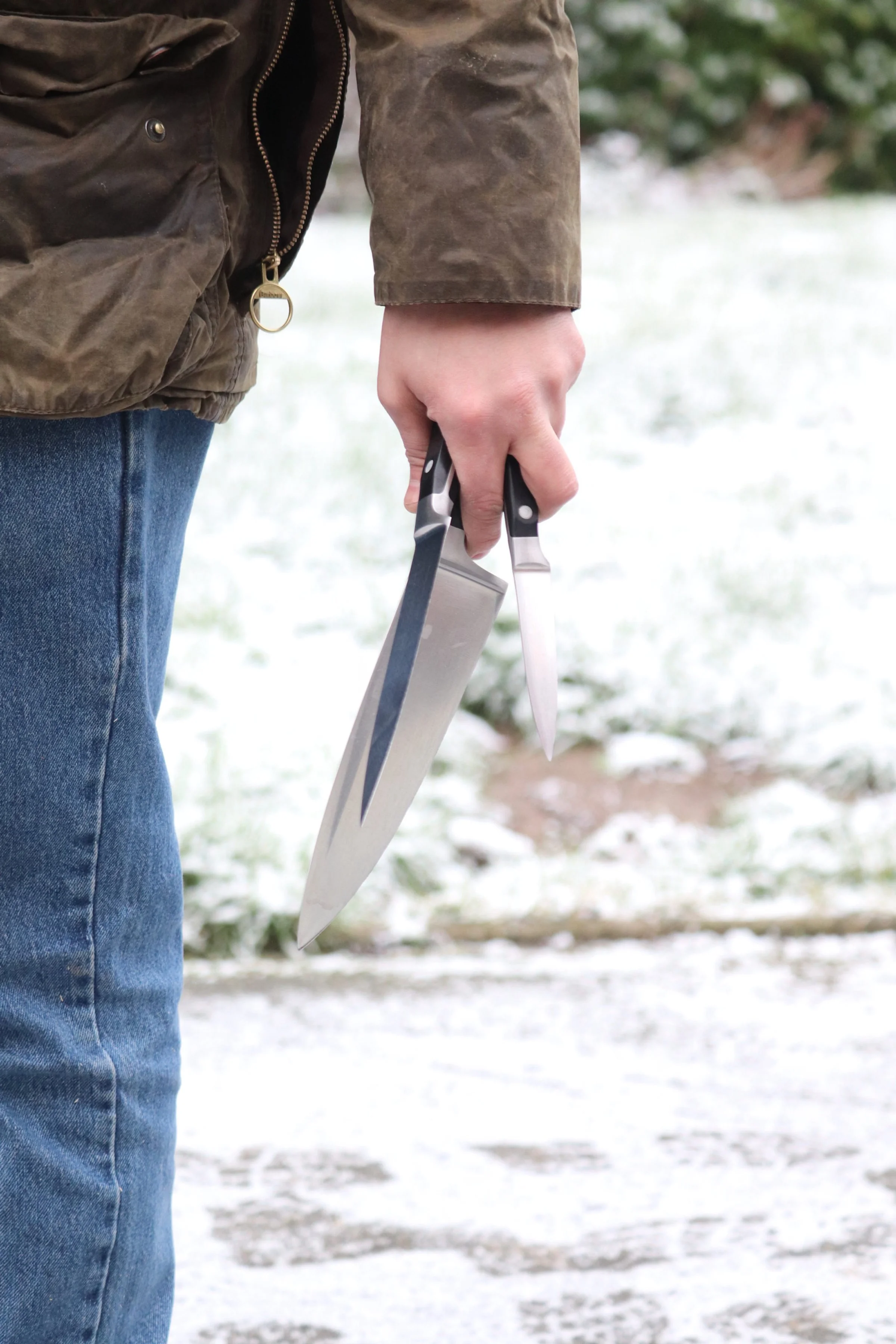 Close-up of a person's hand holding a chef's knife and a paring knife outdoors in a snowy setting, with the person's torso, part of the jacket, and jeans visible.