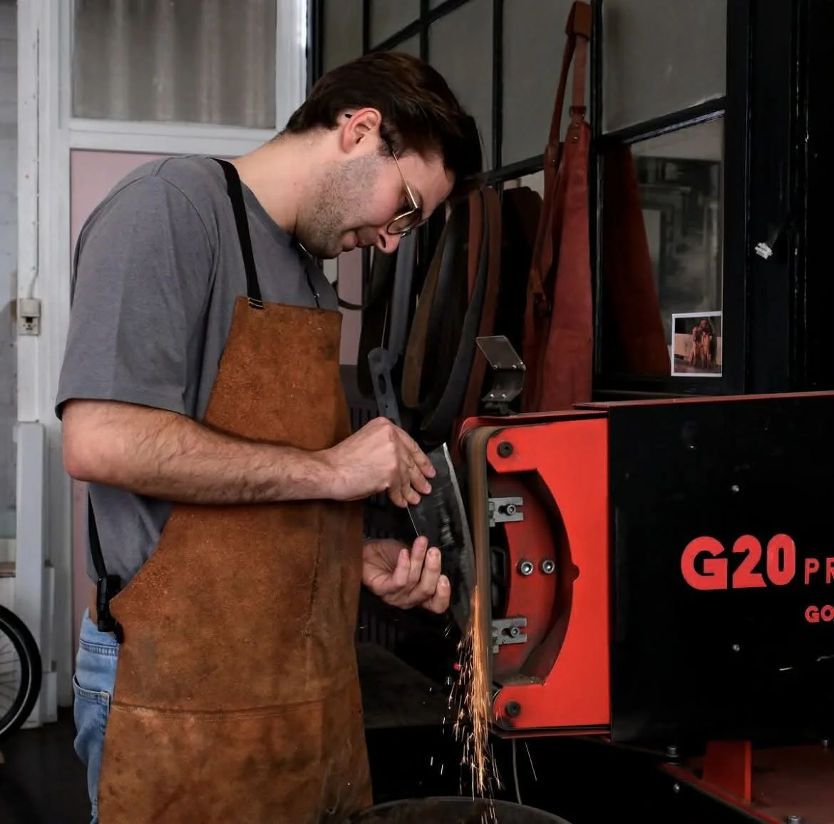 A man wearing glasses, a gray t-shirt, and a brown apron is using a grinding machine, creating sparks, in a workshop.