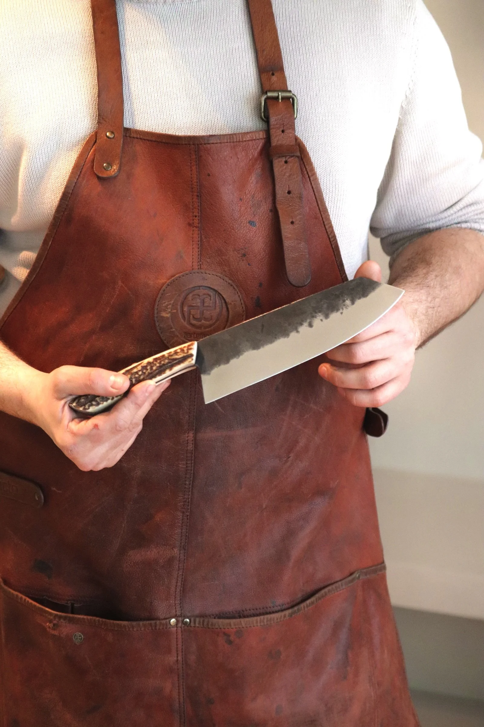 Person wearing a brown leather apron holding a kitchen knife with a textured handle.