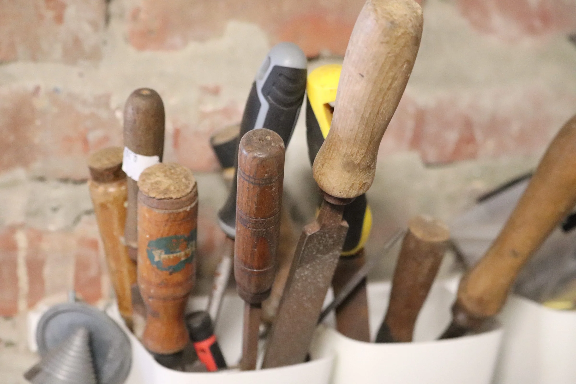 A collection of old and new hand tools including screwdrivers, a hammer, and other woodworking tools, placed in a white container against a brick wall background.