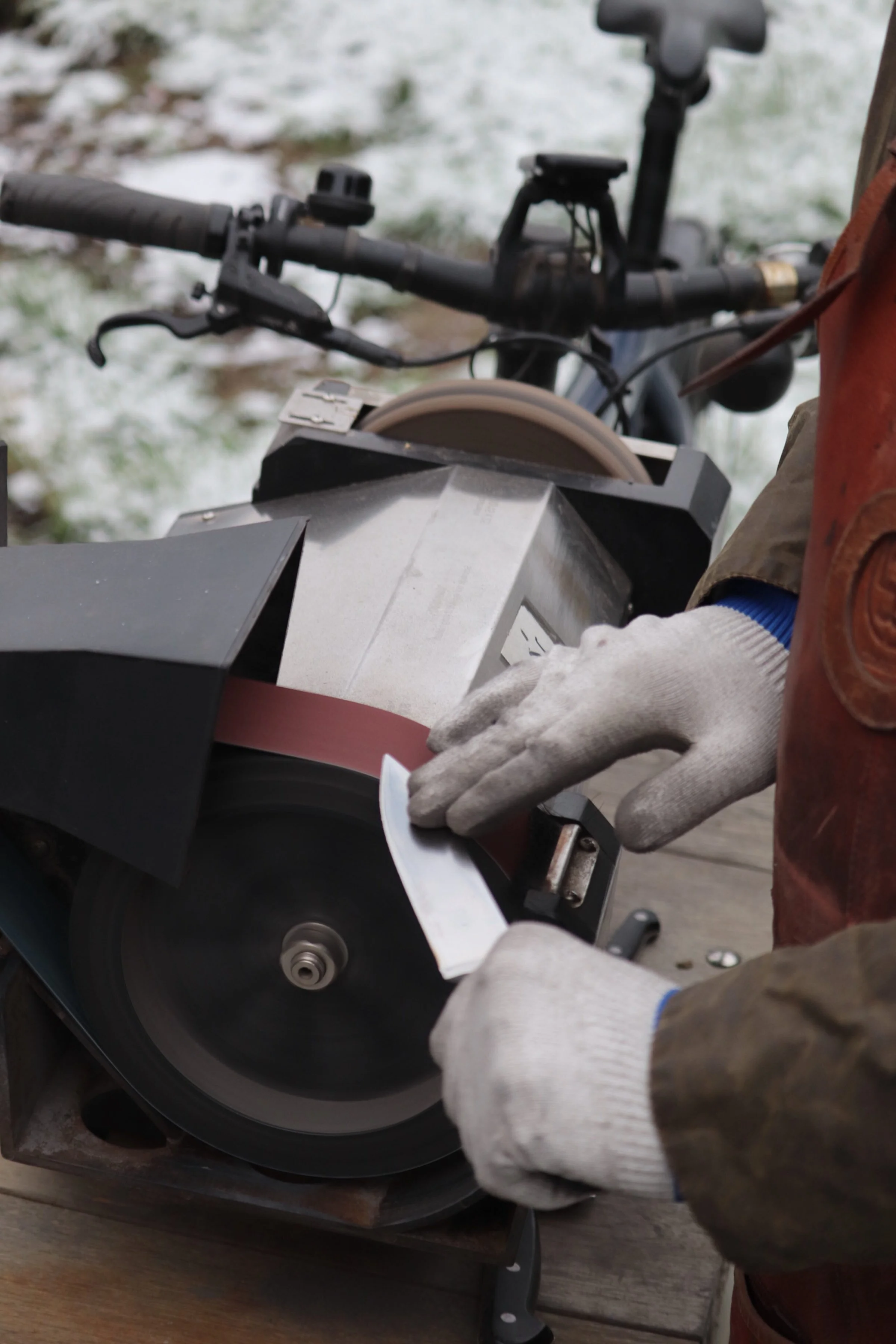 Person wearing gloves using a handheld tool to cut a strip of sanding belt on a bench grinder in an outdoor snowy setting.
