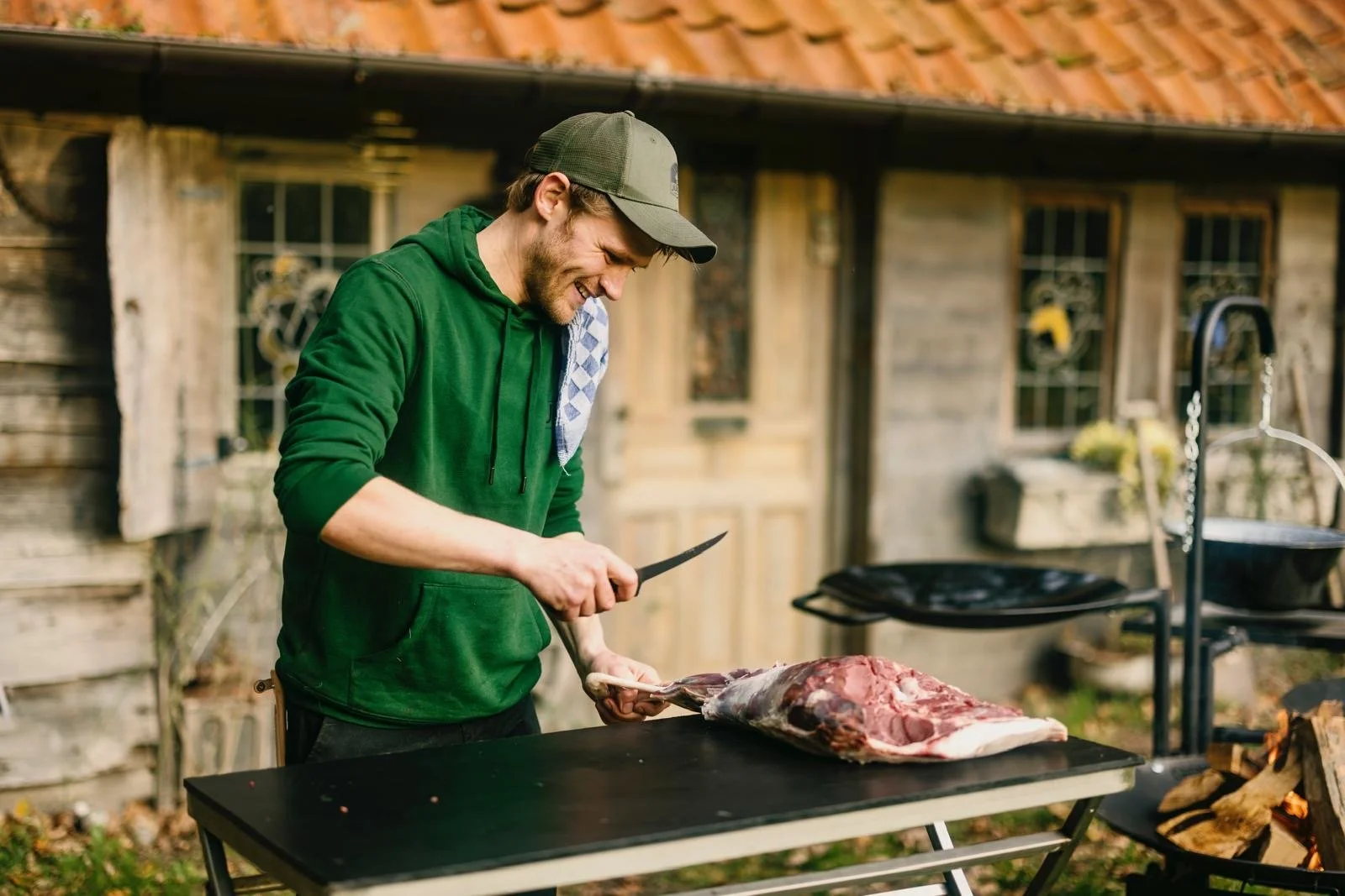 A man in a green hoodie and gray cap preparing meat on a black outdoor grill in a rustic backyard.