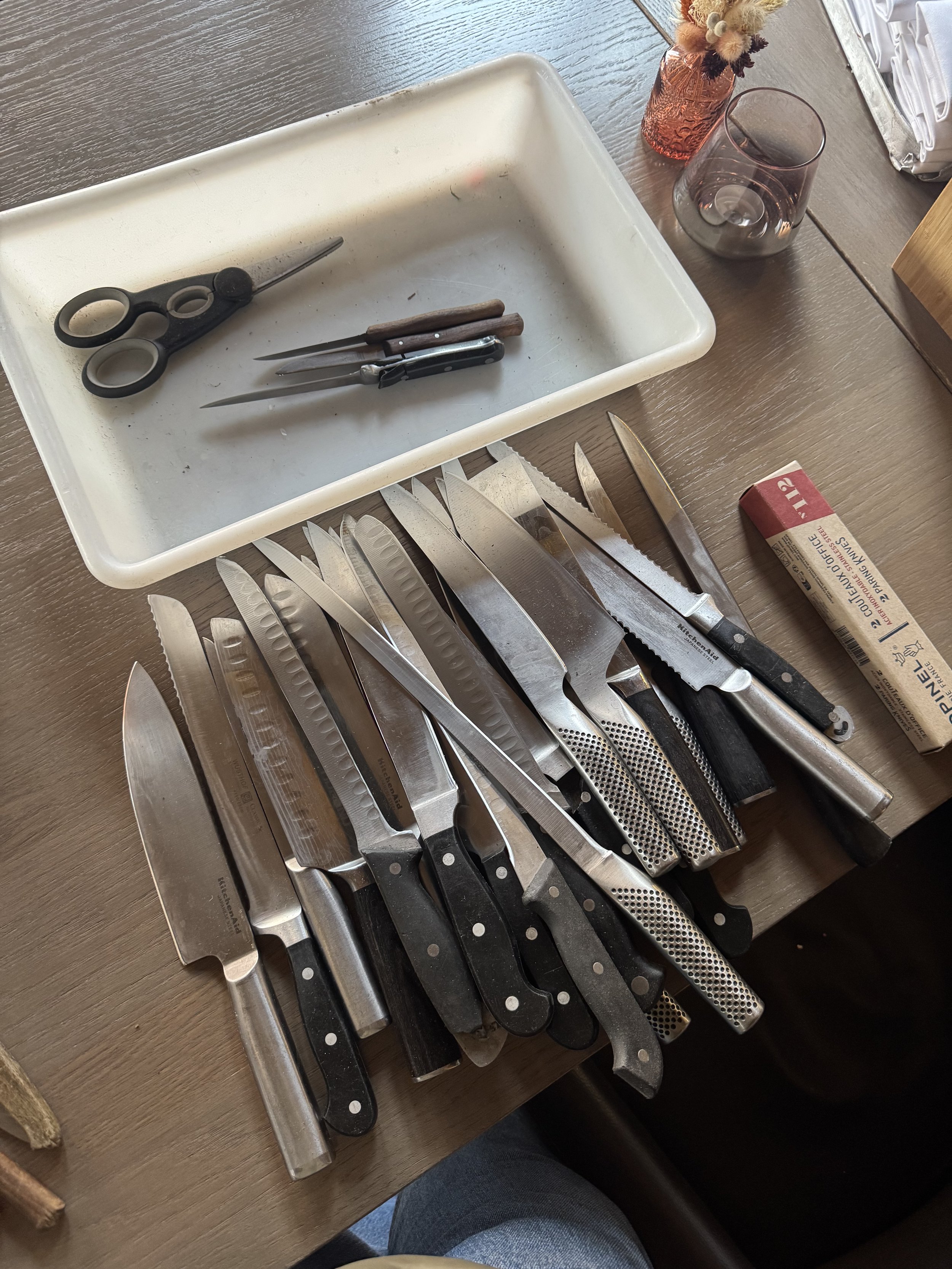 Collection of various kitchen knives, a pair of scissors, and sharpening tools on a wooden table, with a white tray and a pink vase with dried flowers nearby.
