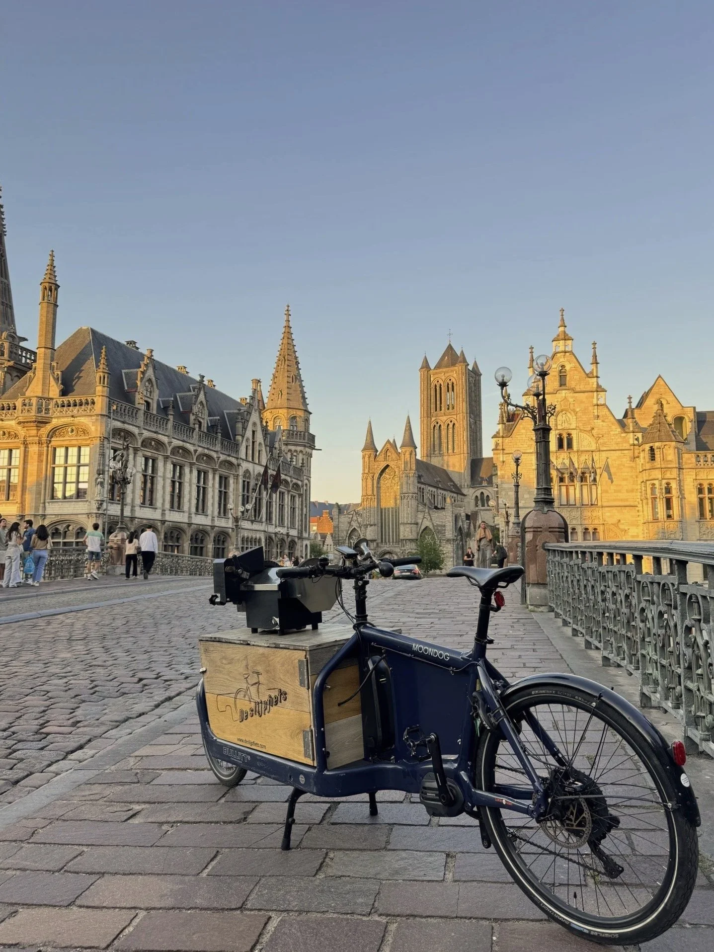 A bicycle with a wooden box on the back, labeled 'De Rippers,' is parked on a cobblestone street in front of historic European-style buildings and churches with tall spires, during sunset.
