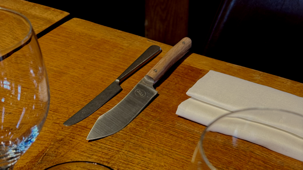 A wooden dining table set with a bread knife with a wooden handle, a butter knife, and a folded white napkin.
