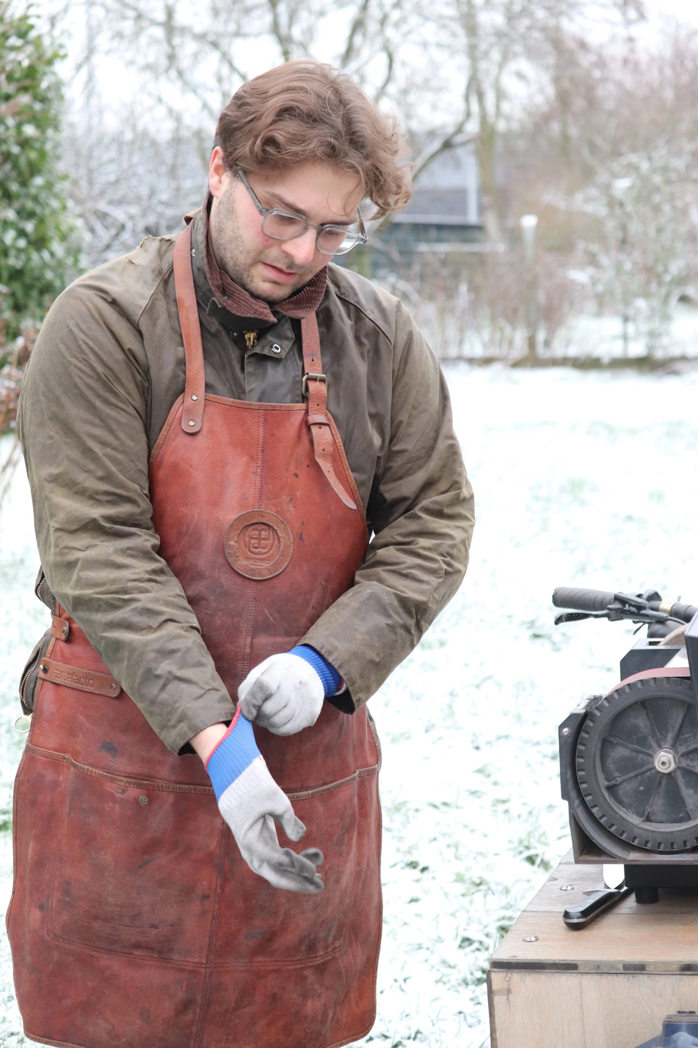 Man wearing glasses and a brown apron, standing outdoors on snowy ground, preparing to cut wood with a saw.