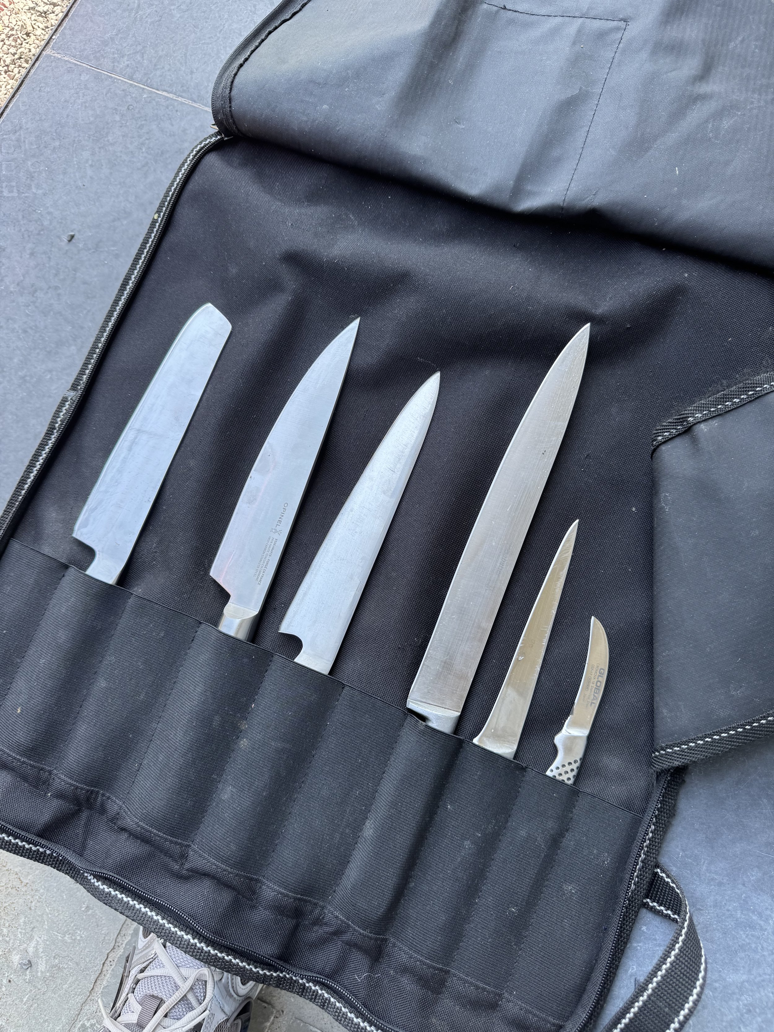 Set of six kitchen knives with varying sizes and shapes resting in a black carrying case on a gray surface.