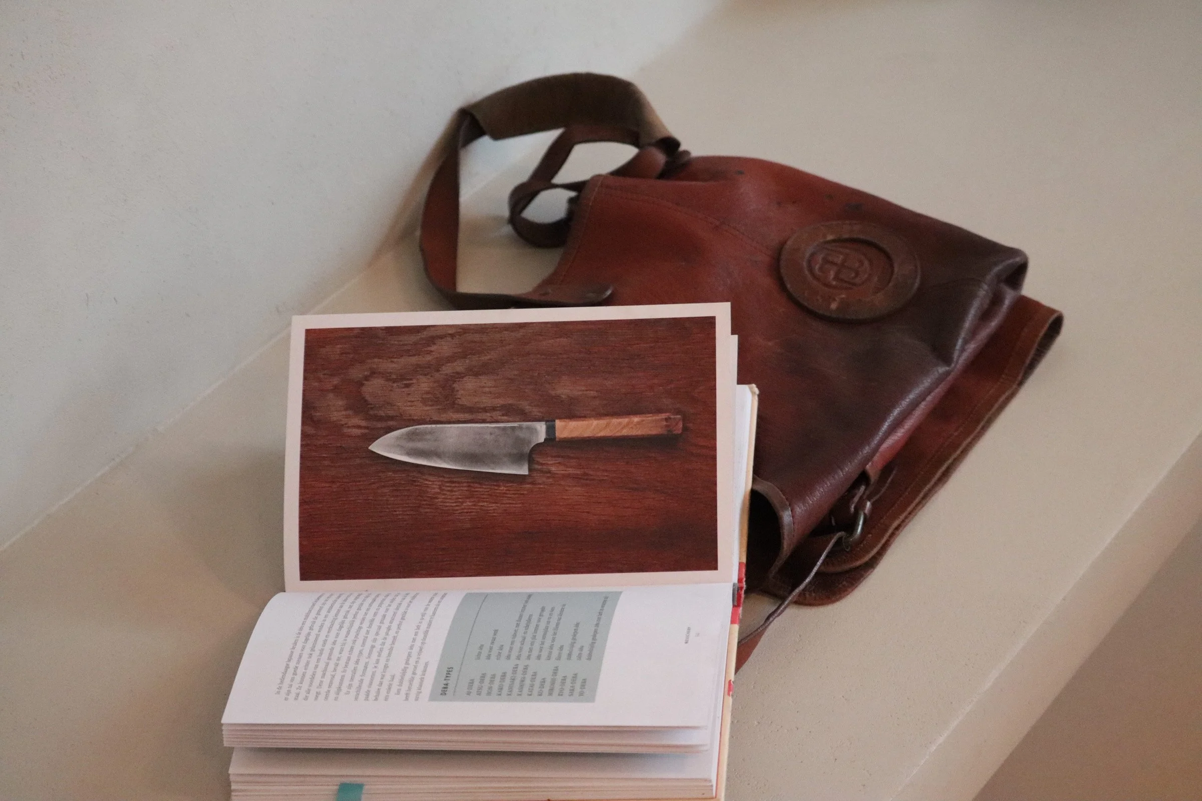 Open book with a photograph of a knife on a wooden surface, placed on a beige surface with a brown leather bag in the background.