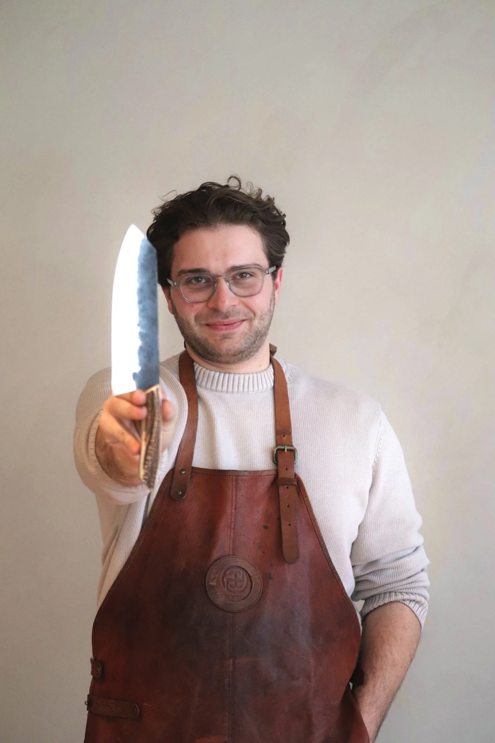 Man wearing glasses and a brown leather apron holding a knife toward the camera, standing against a plain light-colored wall.
