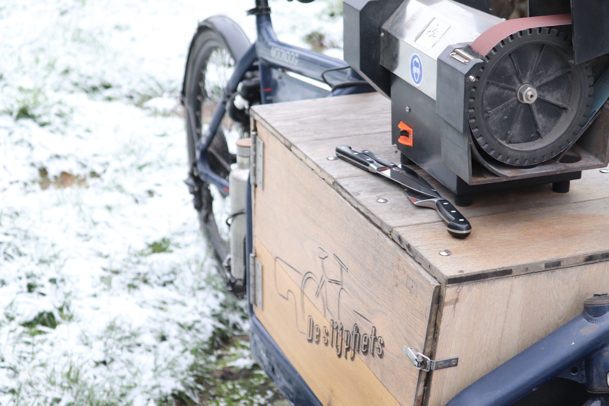 Close-up of a wooden crate with the logo 'delsupets' attached to a motorized vehicle, with a bolt cutter and ice skate blades on top, and a electric scooter in the background on a snow-covered ground.