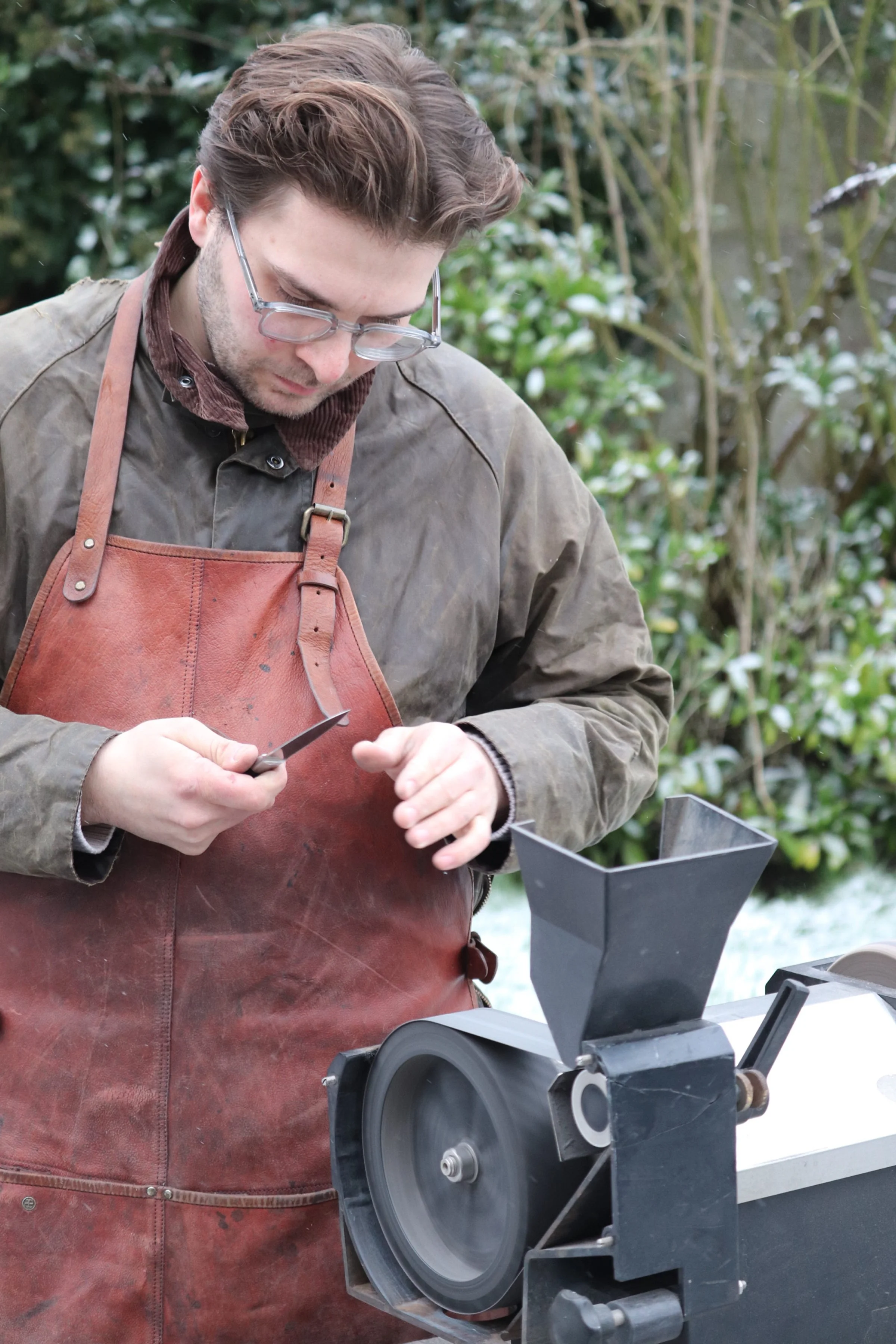 A man wearing glasses and a leather apron using a meat grinder outdoors during winter.