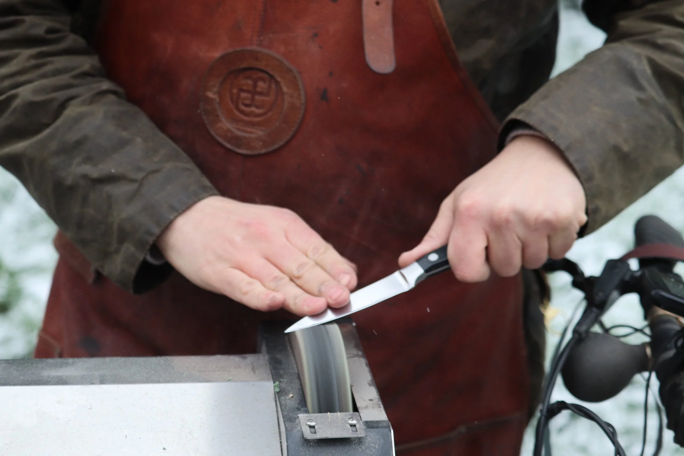 Person wearing a brown leather apron sharpening a knife on a grinding tool.