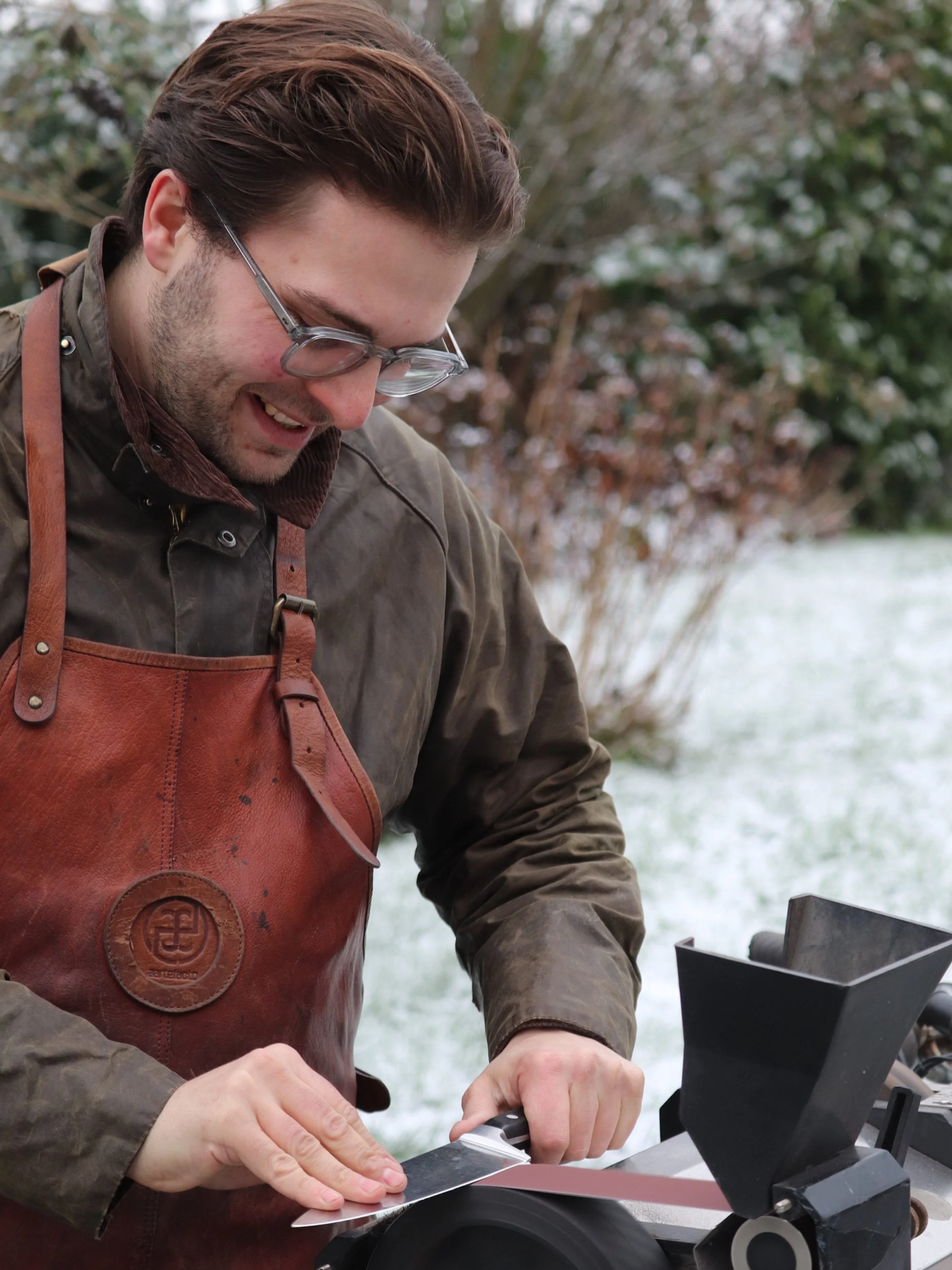 A man wearing glasses, a brown jacket, and a leather apron smiling as he uses a hand saw to cut through a piece of wood outdoors with trees and snow in the background.