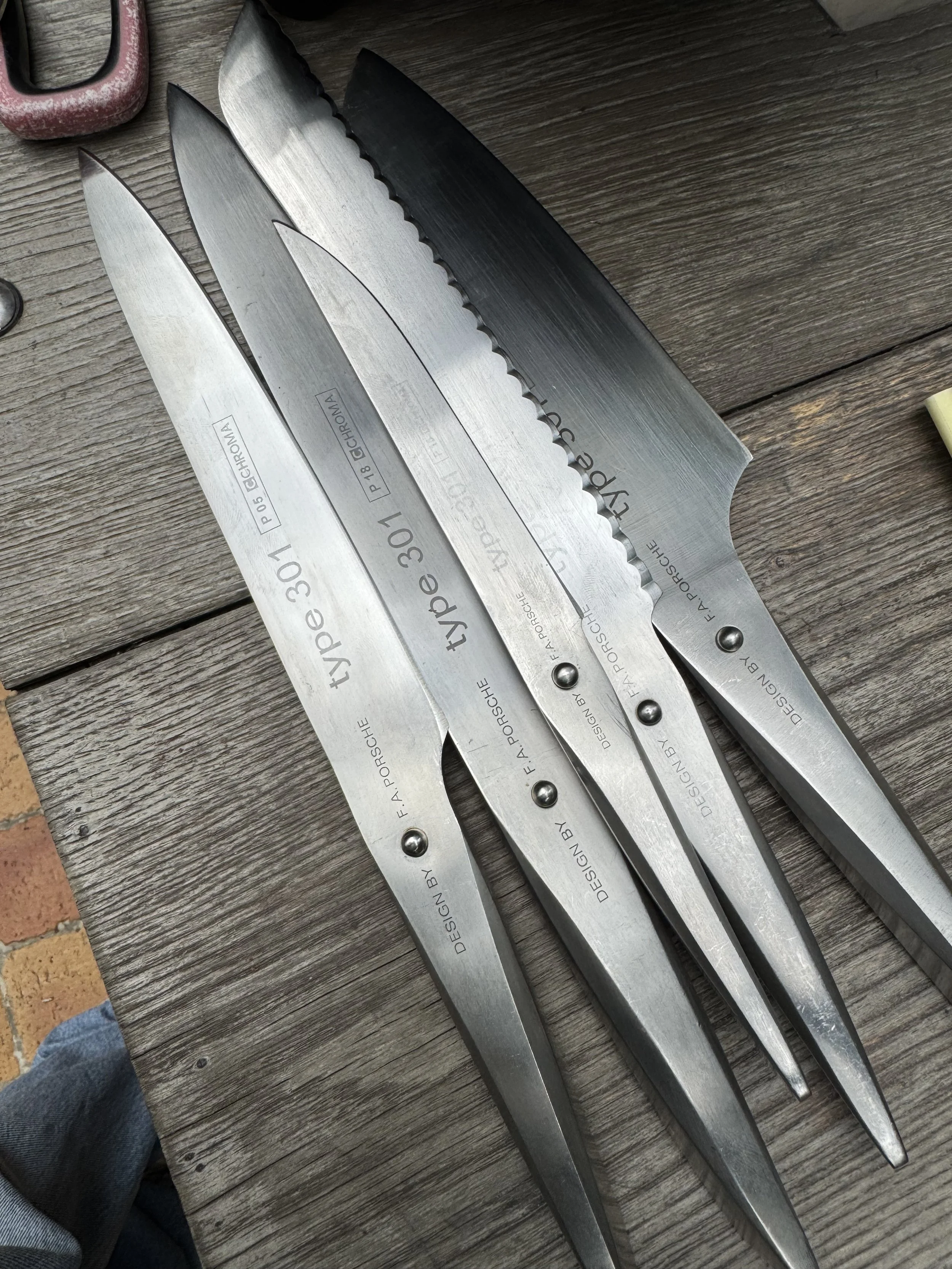 Four kitchen knives with engraved text, placed on a wooden surface. The knives vary in size and shape, with one having a serrated edge.