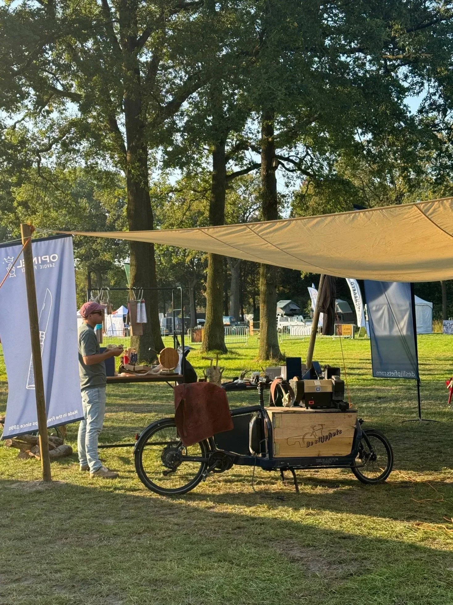 A mobile bicycle food stand at an outdoor event with banners, a person, and large trees in the background.