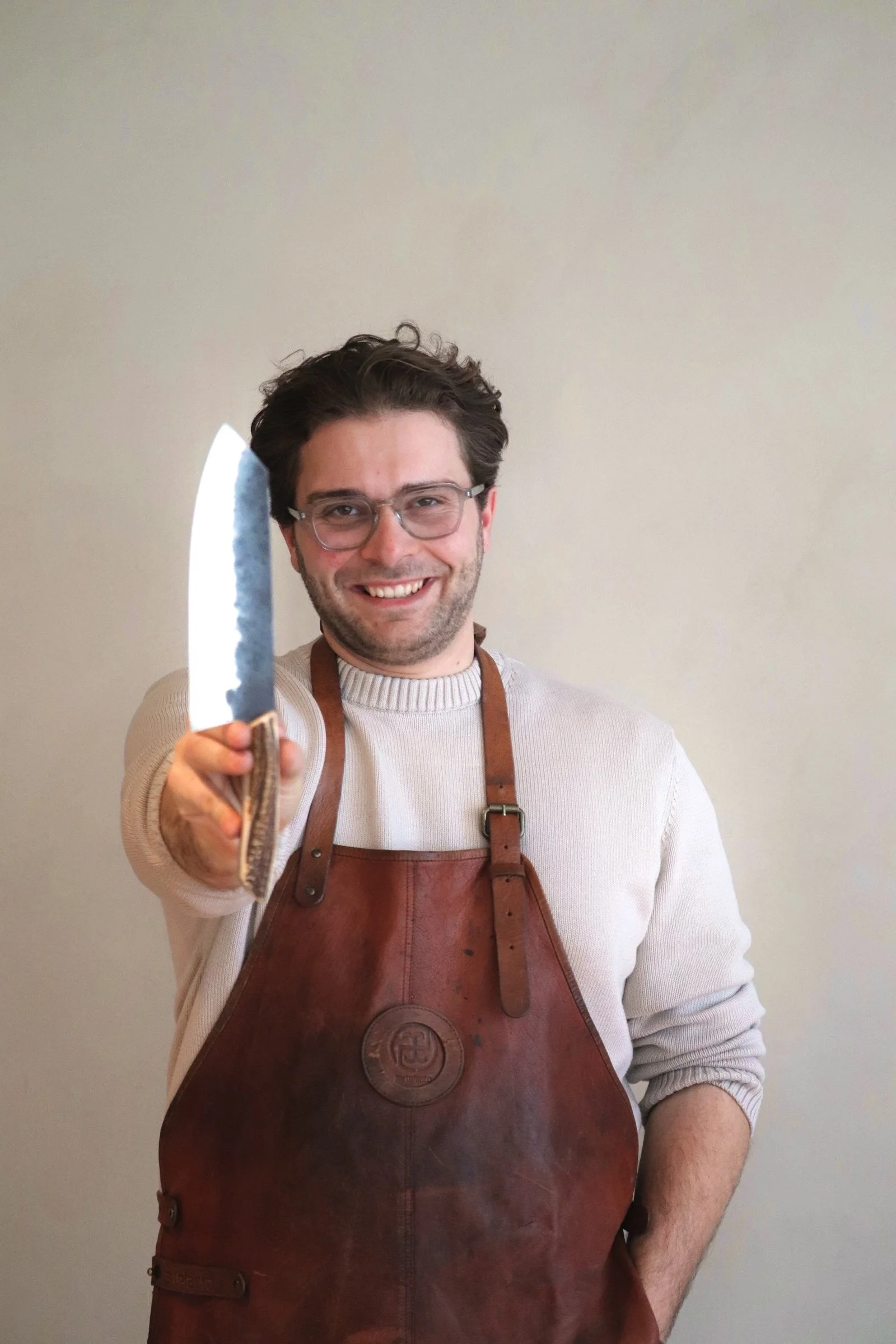 A smiling man with glasses, dark hair, wearing a beige sweater and a leather apron, holding a kitchen knife towards the camera.