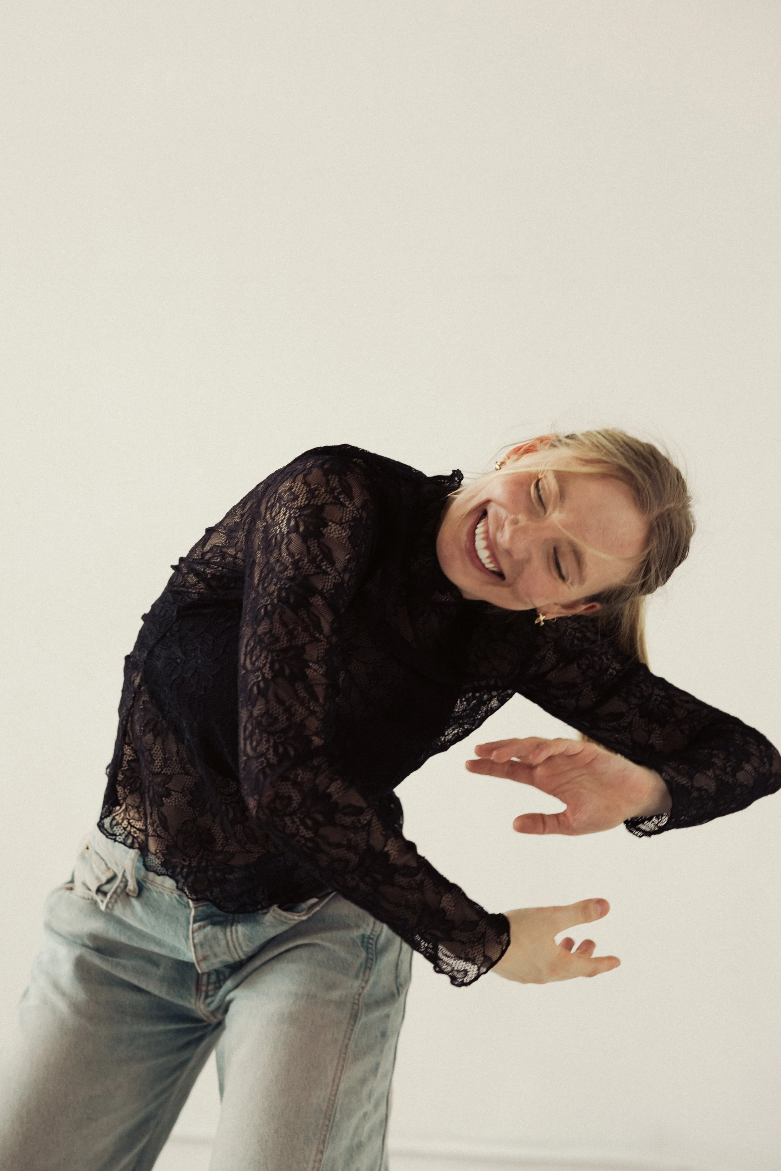 A woman laughing and smiling while wearing a black lace top and light-colored jeans against a plain white background.