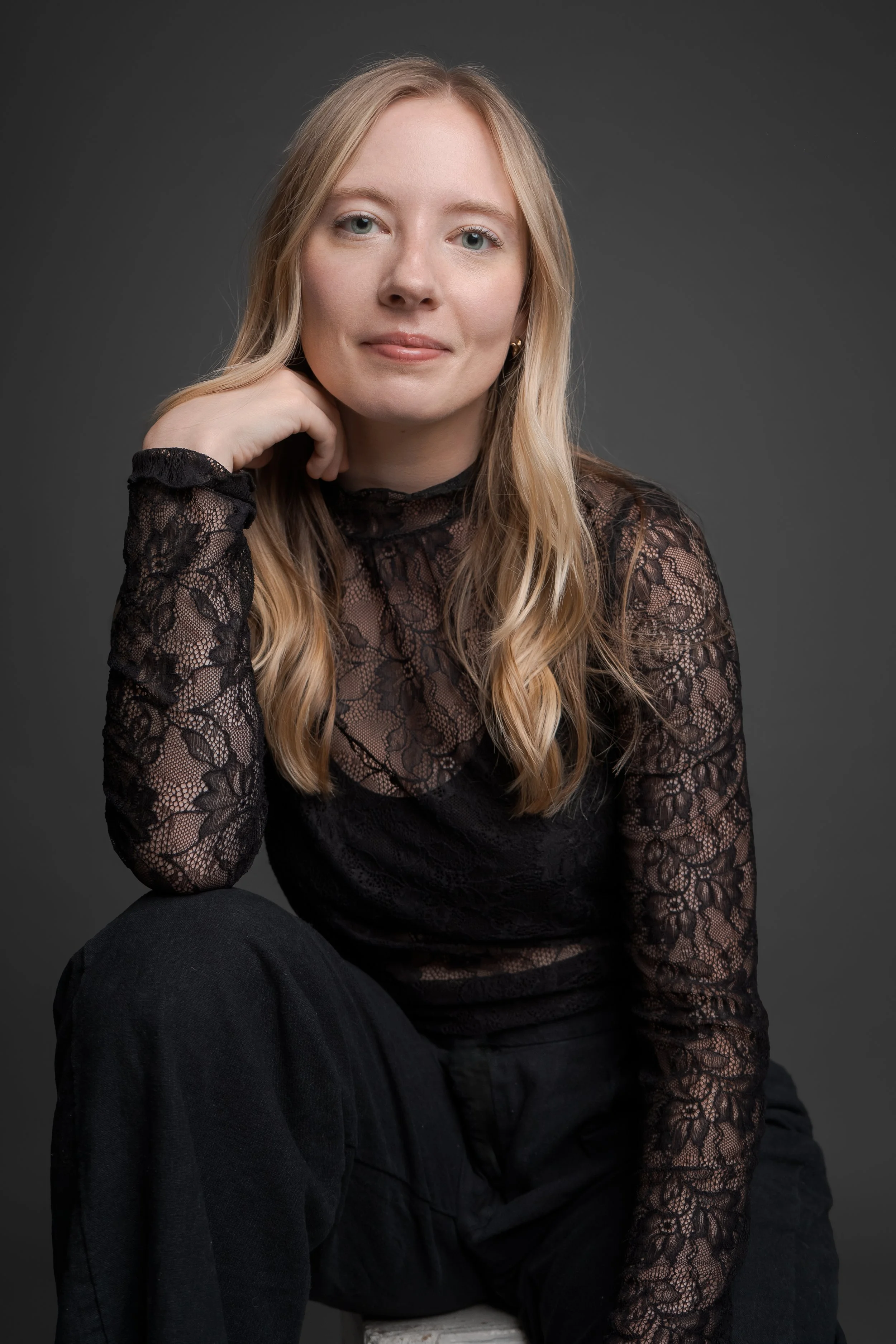 Portrait of a young woman with long blonde hair, wearing a black lace top and black pants, sitting against a dark grey background.