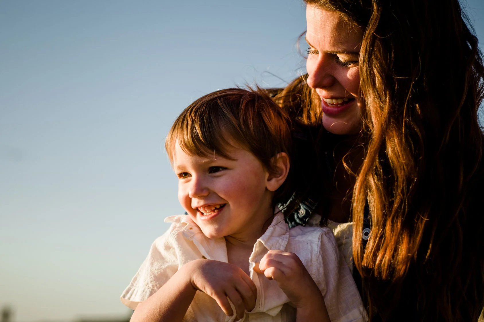 Femme et jeune garçon souriants en extérieur, regardant vers la gauche, avec un ciel bleu en arrière-plan.