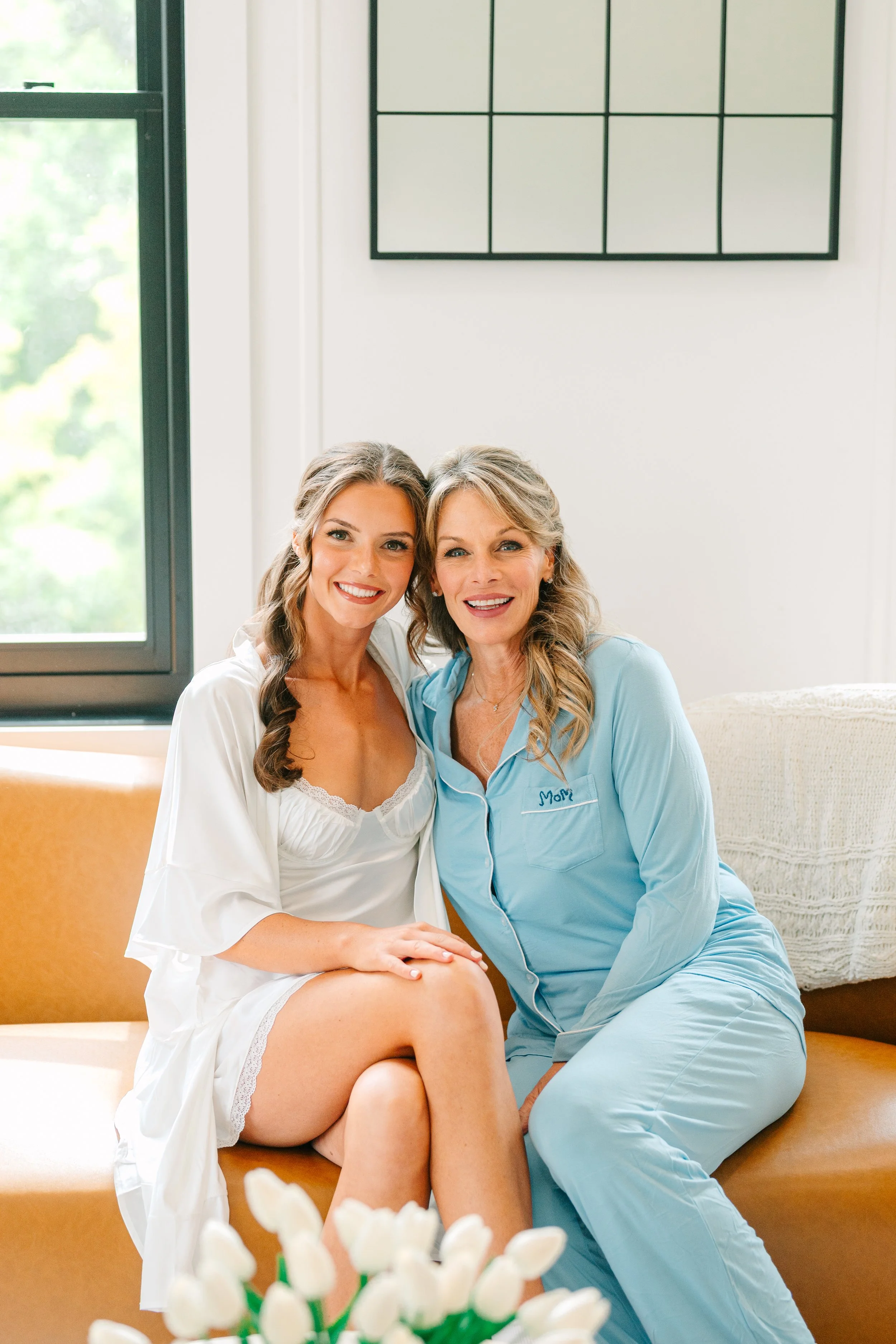 A woman in a white nightgown and a woman in blue pajamas sitting on a yellow couch, smiling, with a window in the background. Wedding Cleveland Ohio Planner