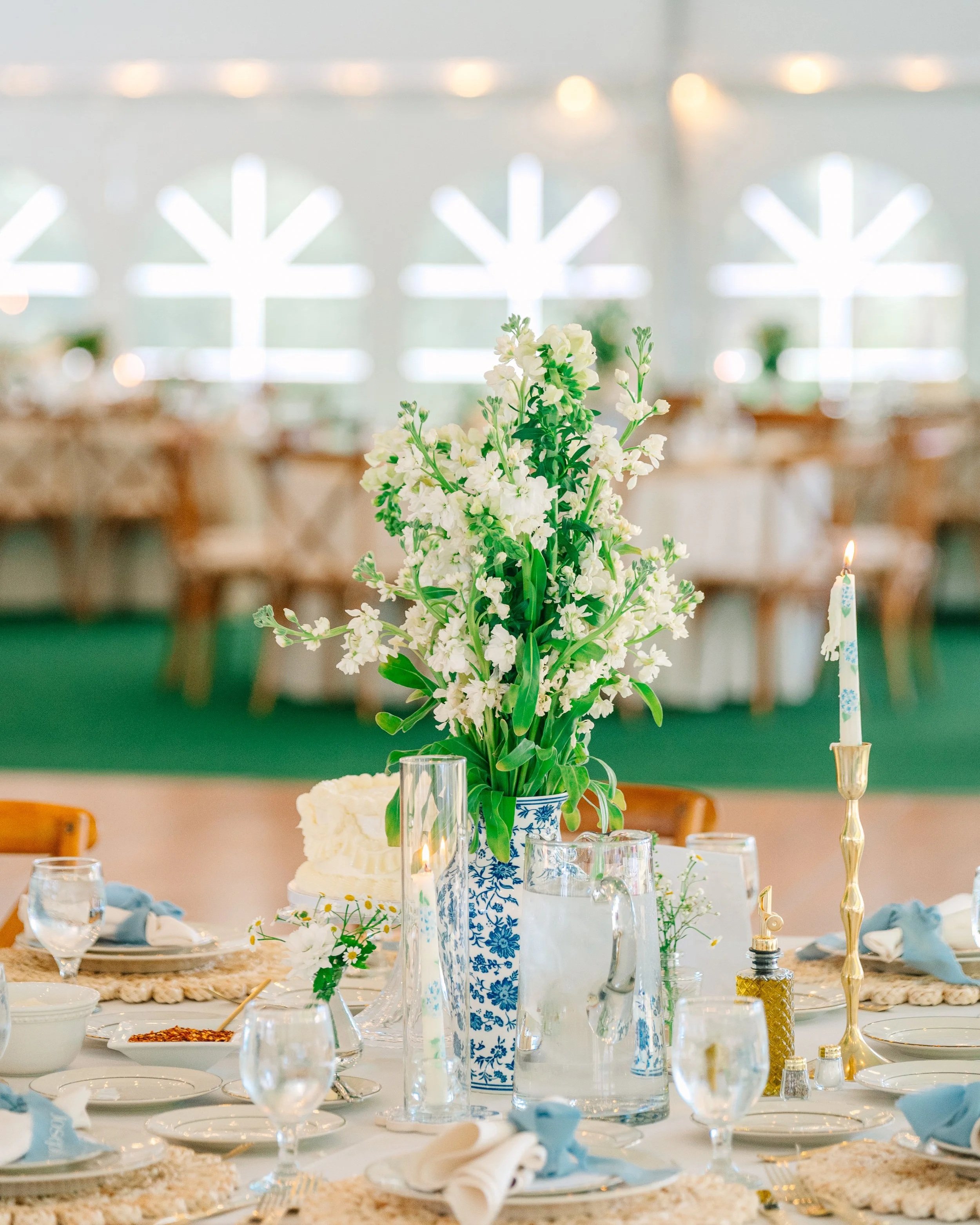 A table set for a formal event with a large white floral arrangement in a blue and white vase, tall candles, and various glassware and plates in a decorated room.