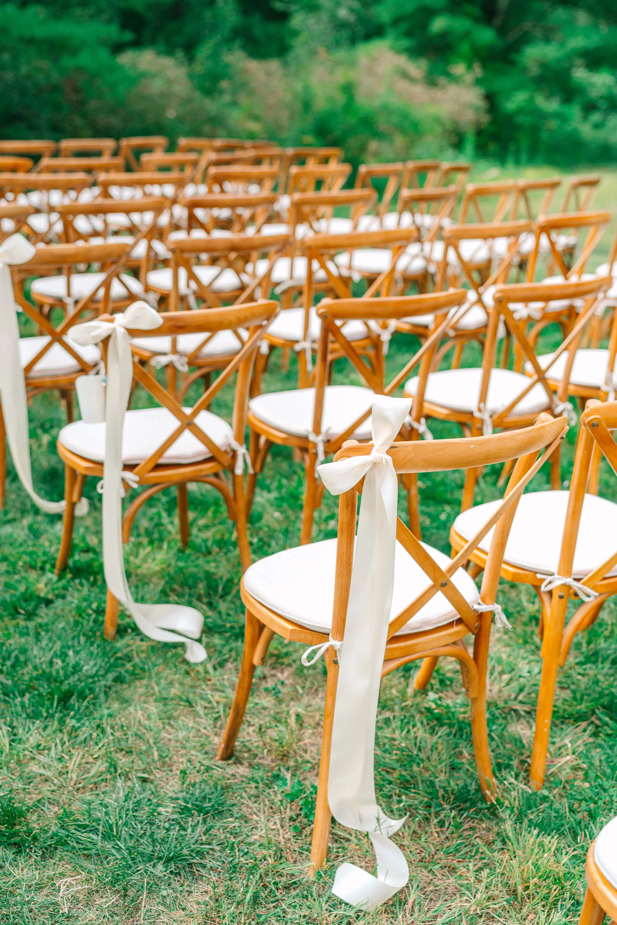 Multiple wooden chairs with white cushions and tied white ribbons are arranged outdoors on grassy ground, likely set up for a wedding or special event.