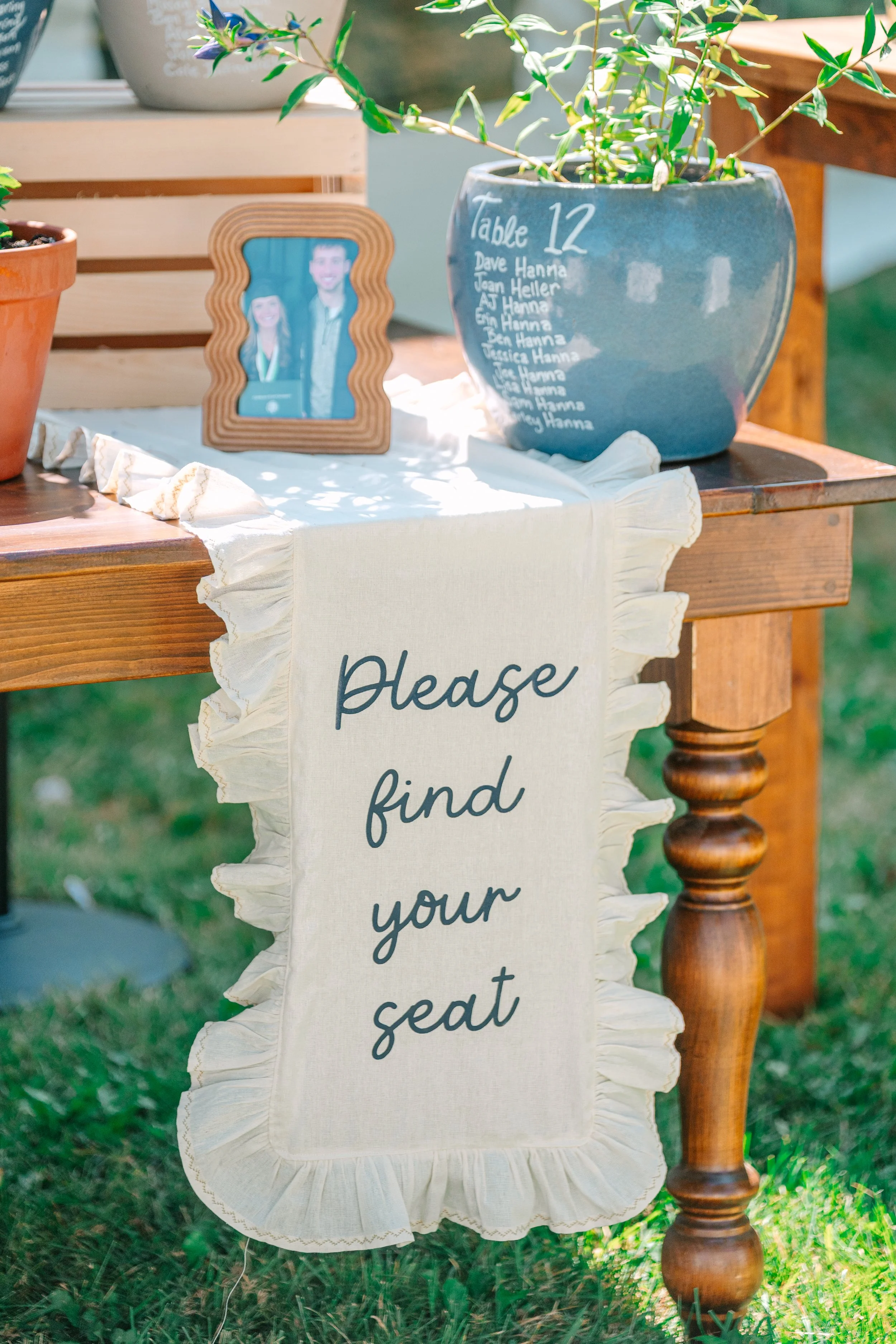 A table at an outdoor event with a sign that reads 'Please find your seat,' a picture frame of two people, a chalkboard table number with a list of names, and flower pots on a wooden table.