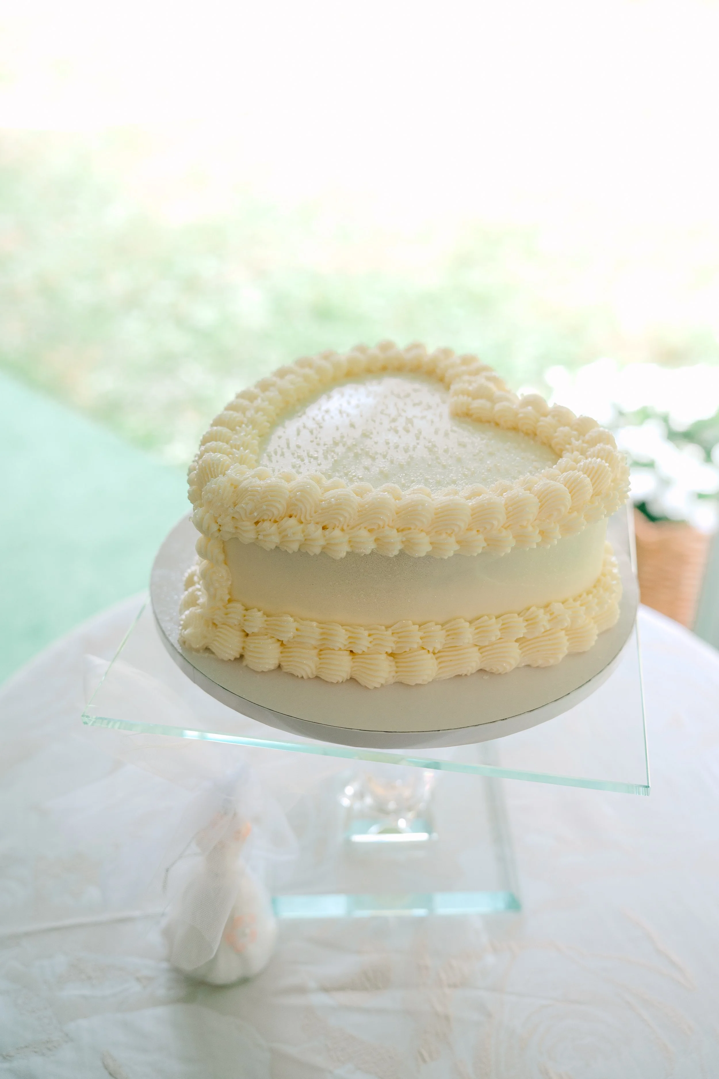 A heart-shaped white wedding cake with piped cream borders on a glass stand, set against a bright, blurred background.