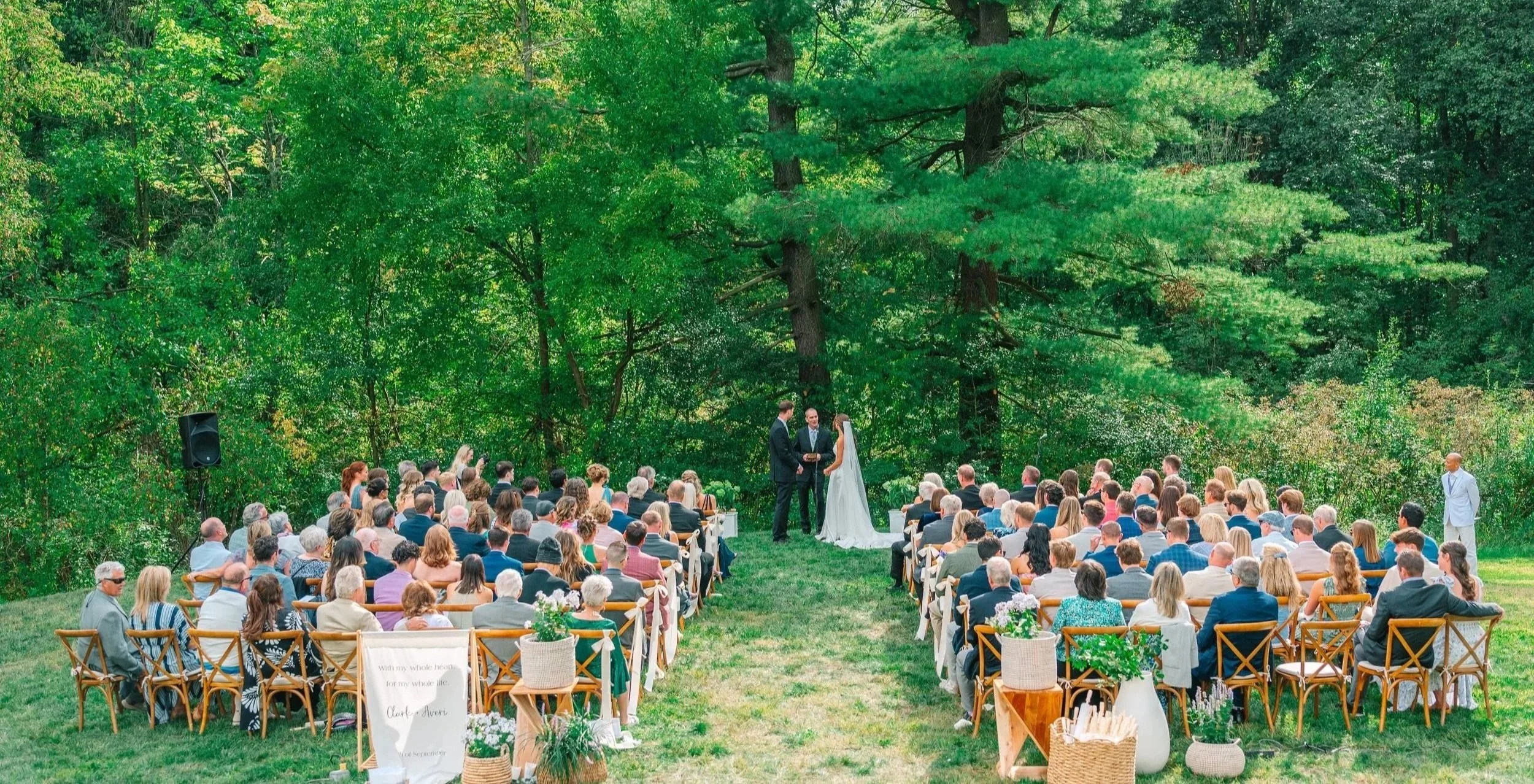 Outdoor wedding ceremony in a lush green forest with seated guests, a bride and groom at the altar, and officiant. Akron Ohio