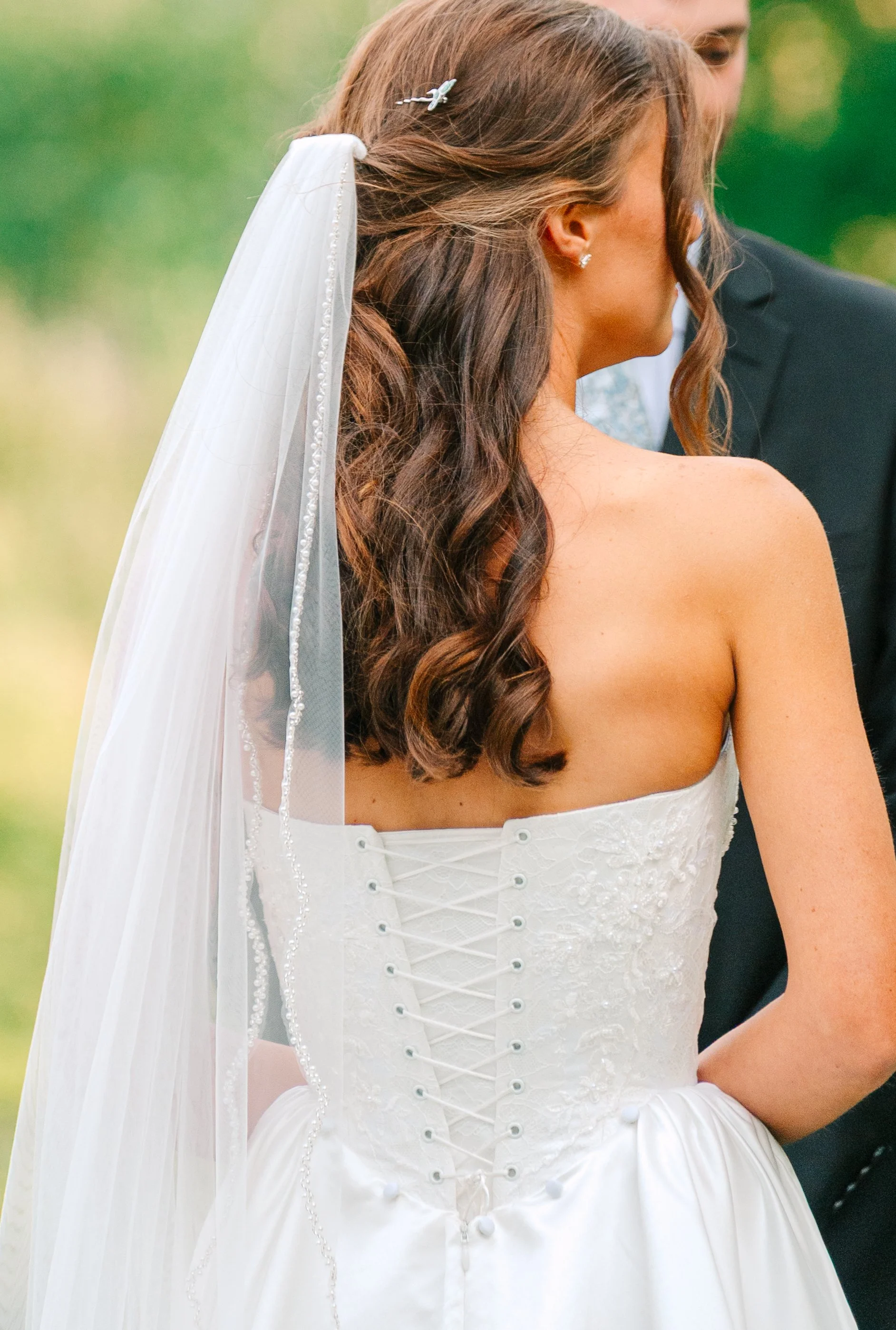Bride in a strapless white wedding gown with lace-up back, long brown hair with curls, and a veil, facing a groom in a black suit, outdoors with green blurred background. Akron Ohio wedding planner