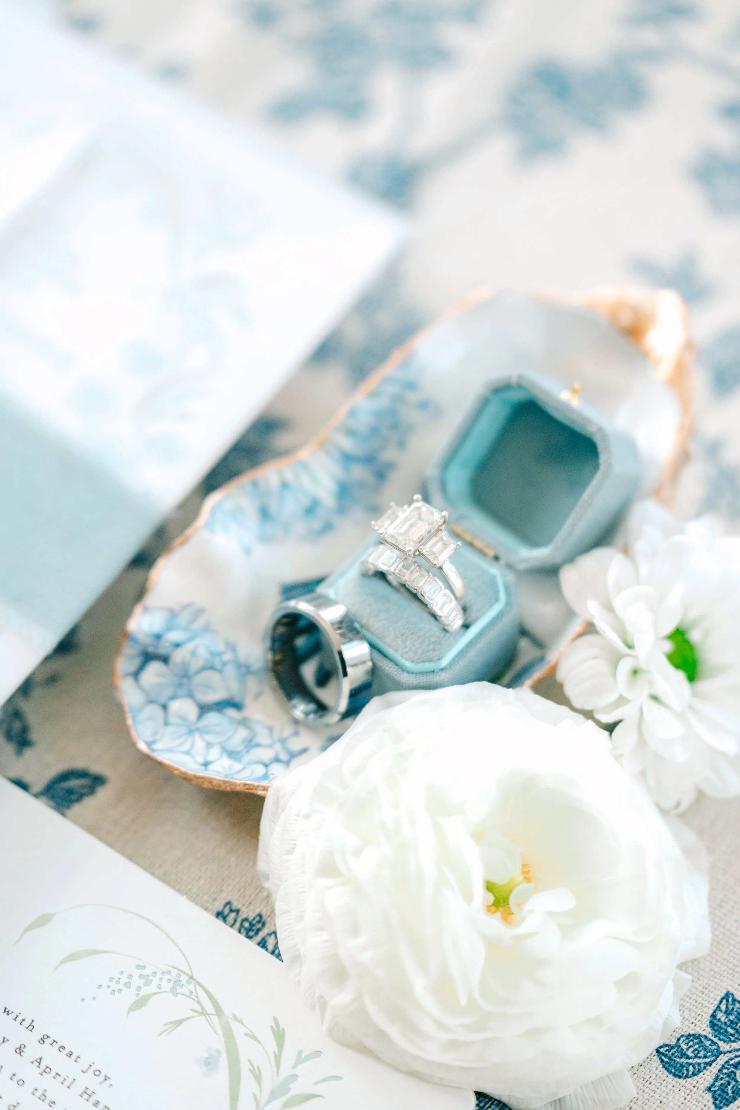 A set of wedding rings in a decorative blue jewelry box, surrounded by white flowers and a partially visible invitation card.