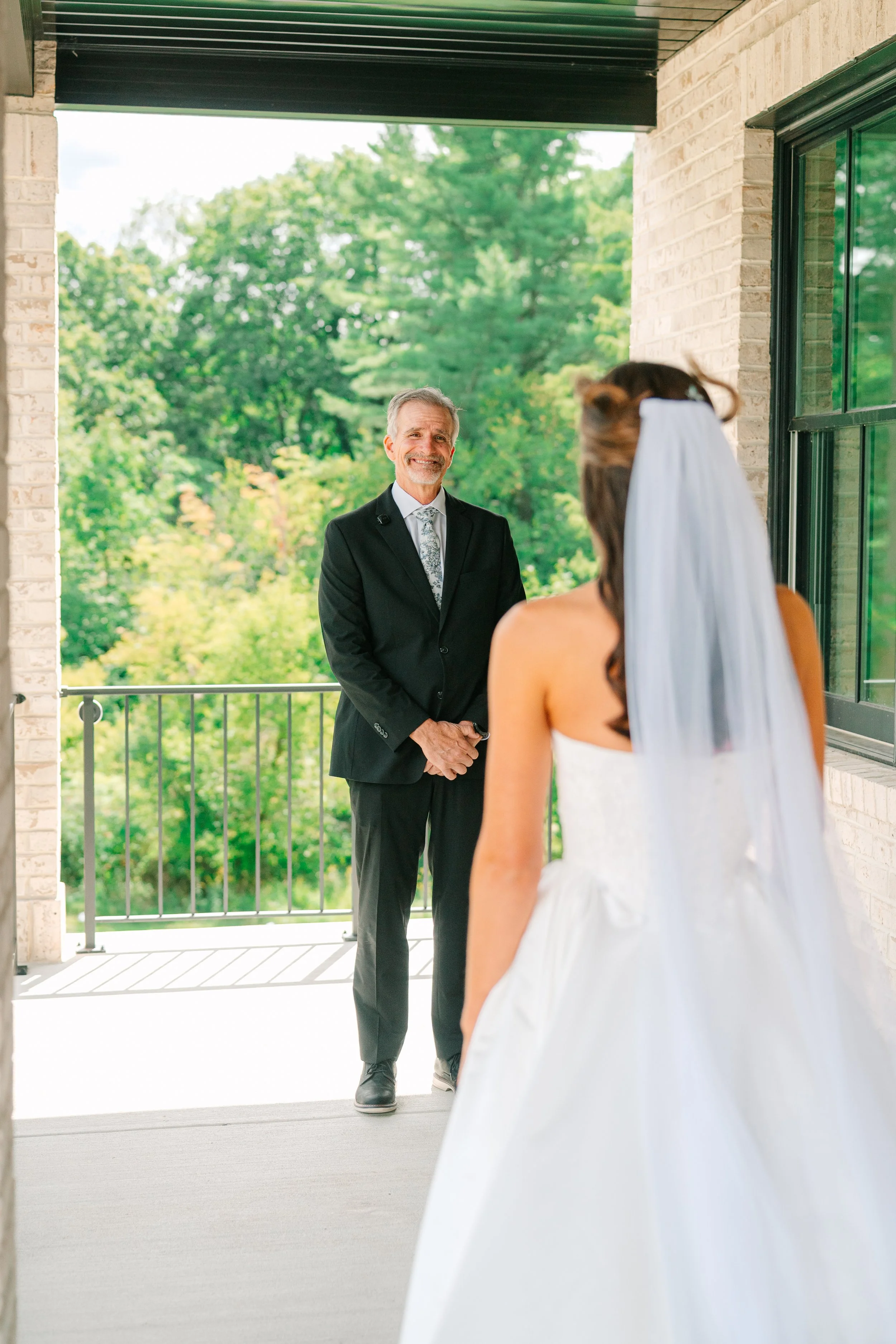 A bride in a white wedding dress and veil standing on a balcony facing an officiant, a man in a black suit with a patterned tie, outdoors with green trees in the background.