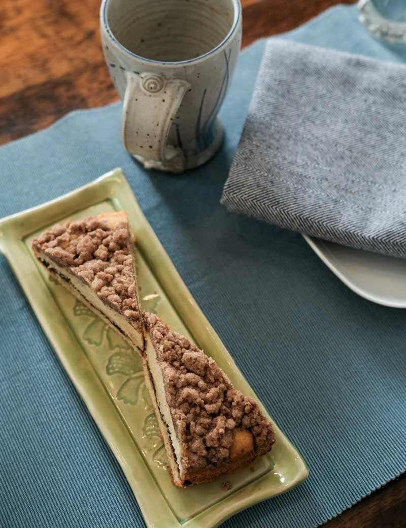 A slice of crumb cake on a decorative green plate, a ceramic mug, a gray cloth napkin, and a partially visible white plate on a blue tablecloth.
