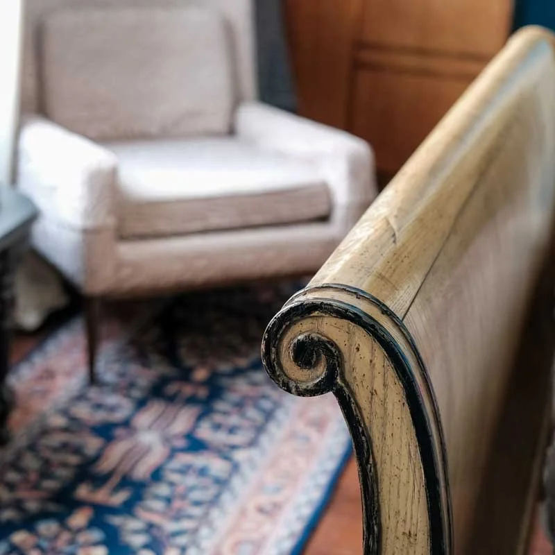 Close-up of a worn wooden armrest with black trim, part of a vintage bench or sofa, in a cozy living room with an armchair and a colorful patterned rug.