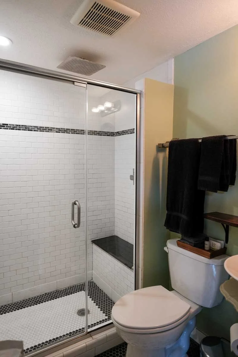 A bathroom with a glass shower enclosure, black and white tiled floor, a white toilet with a wooden tray on top, and black towels hanging on a rack above the toilet.