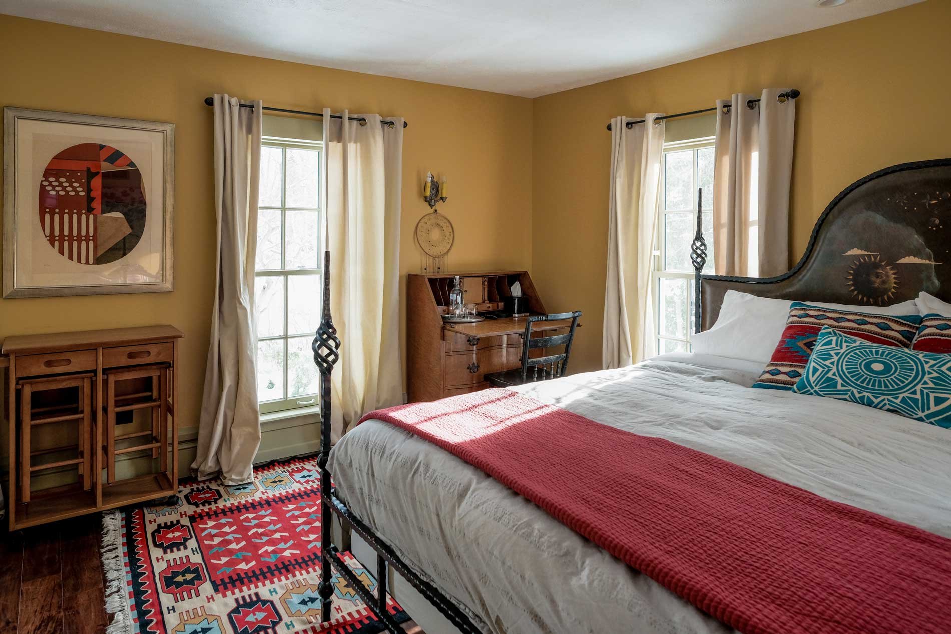 A cozy bedroom with yellow walls and wooden furniture, including a bed with patterned pillows and a red throw, a small wooden dresser, and a vintage writing desk by the window. The room features two large windows with white curtains and a colorful geometric rug on a dark wood floor.