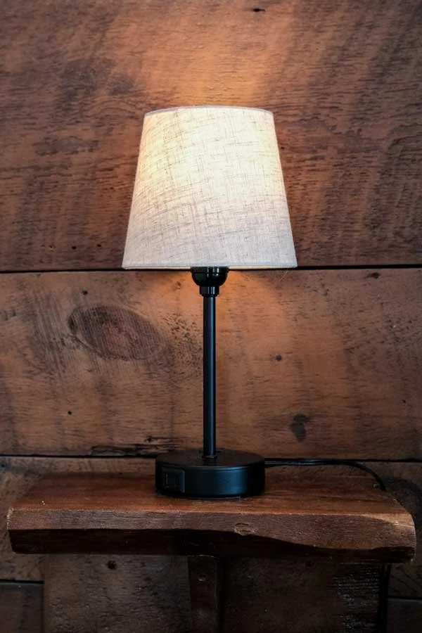 A table lamp with a beige fabric shade and black base, placed on a wooden surface against a wooden wall background.
